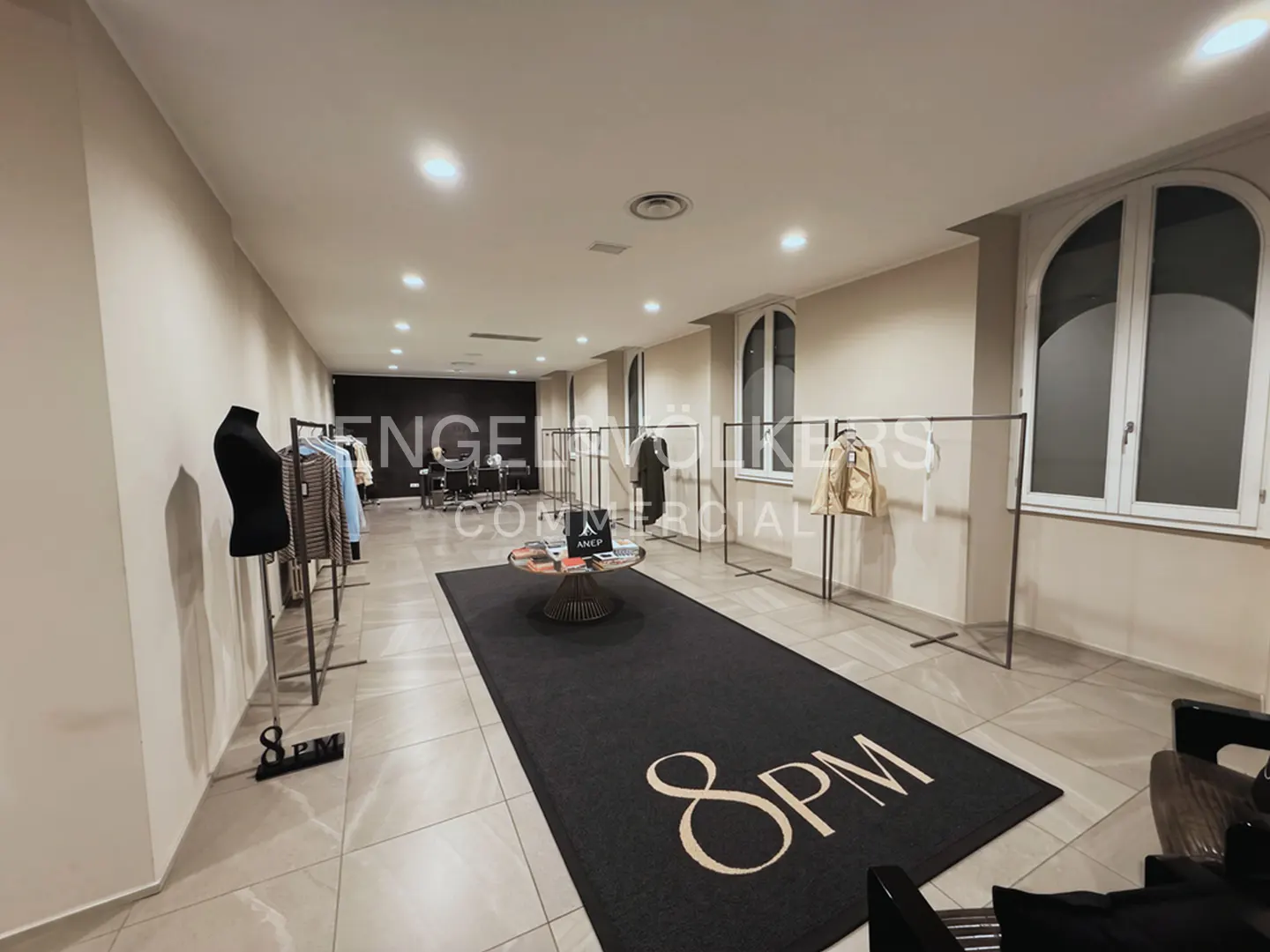 Modern clothing showroom with a black "8PM" logo rug, clothing racks, a mannequin, and arched windows in a bright space.