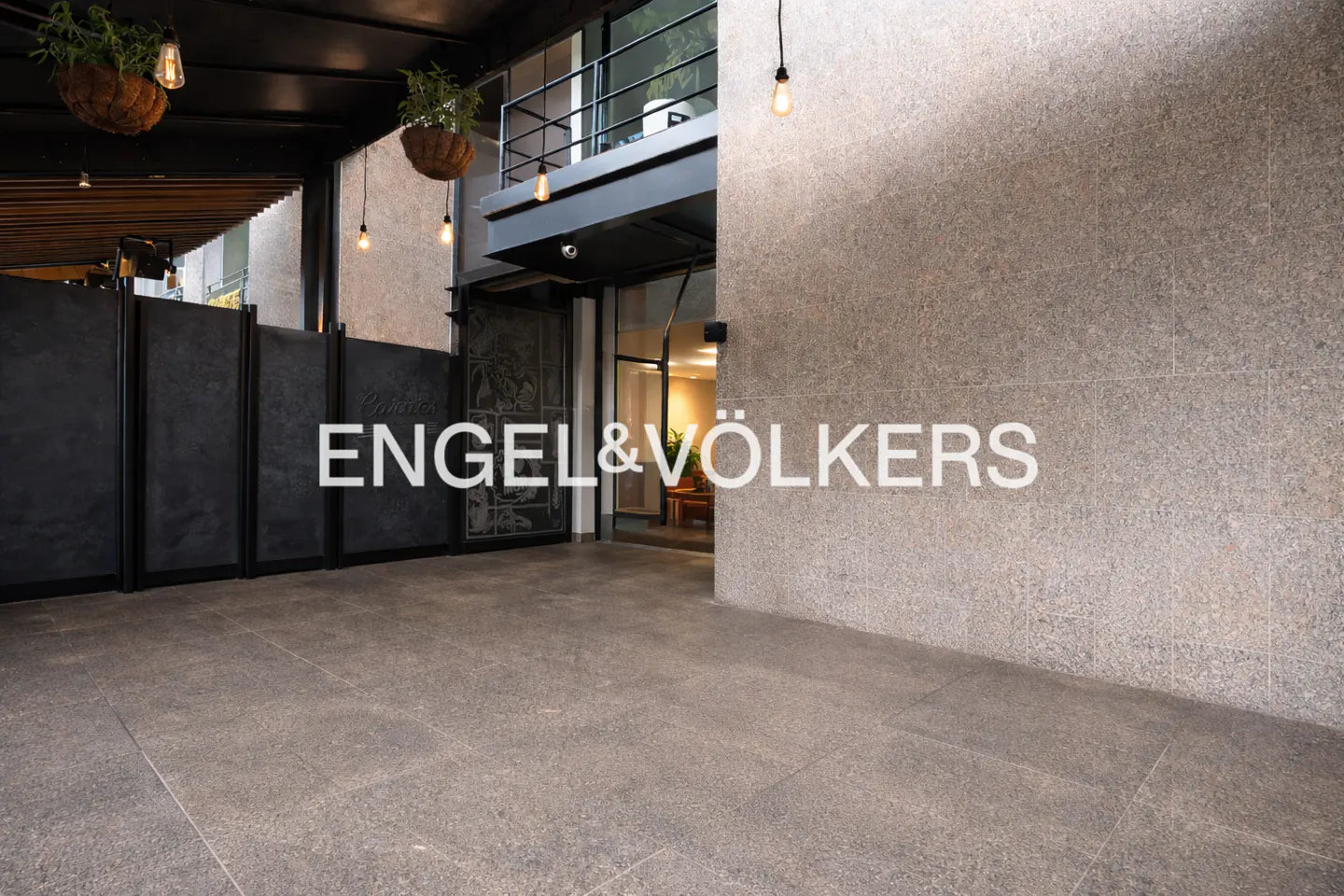 Engel & Völkers office entrance with gray stone walls and floor. Hanging plants and lights decorate the black metal awning.