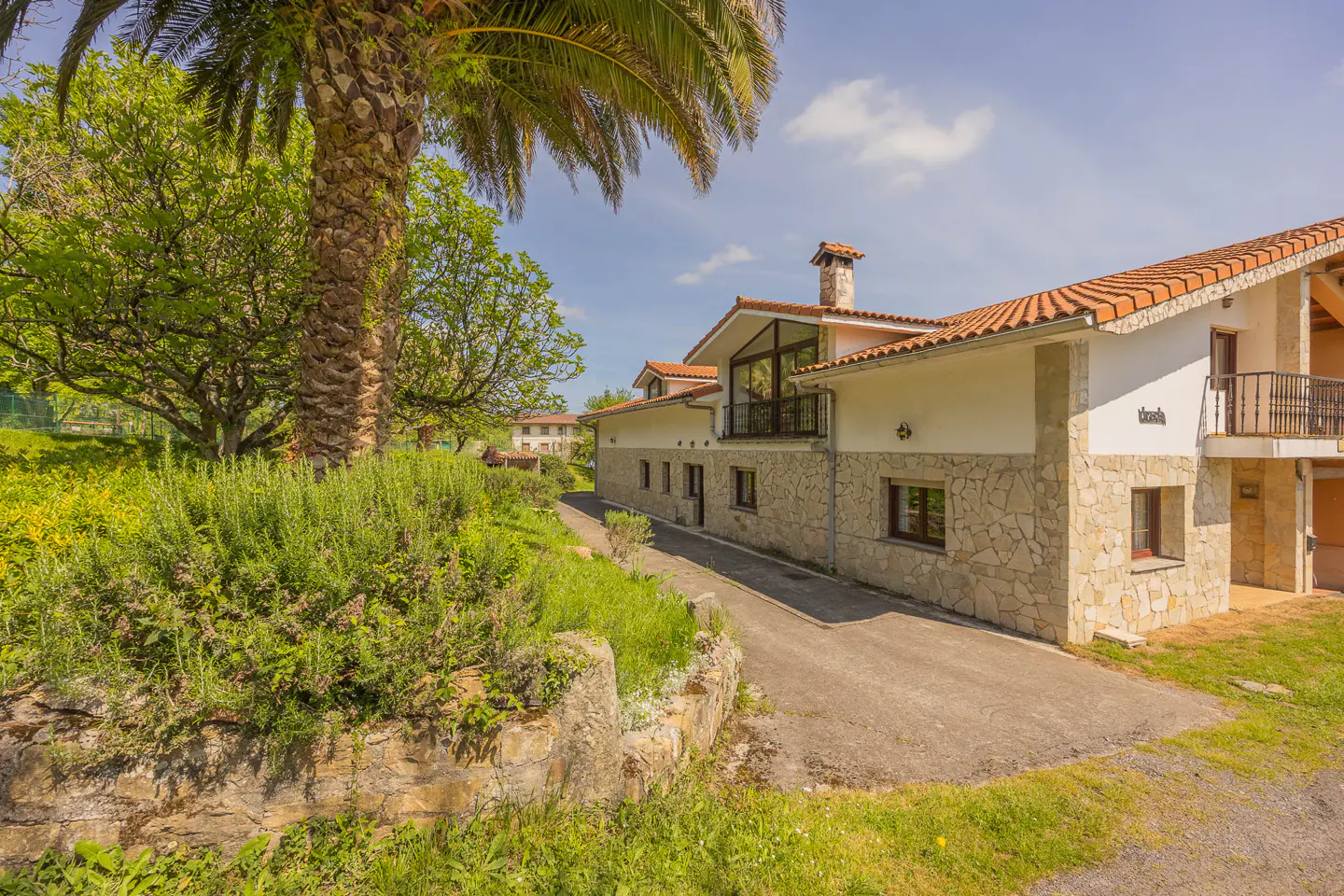 Exterior view of a two-story house with a stone facade and red tile roof, surrounded by green trees and a palm tree.