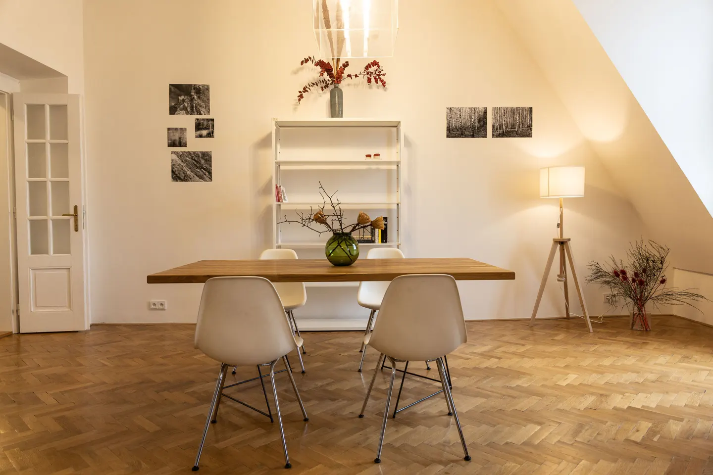 A dining room with a wooden table, white chairs, and herringbone wood floors. A white bookshelf and artwork decorate the white walls.