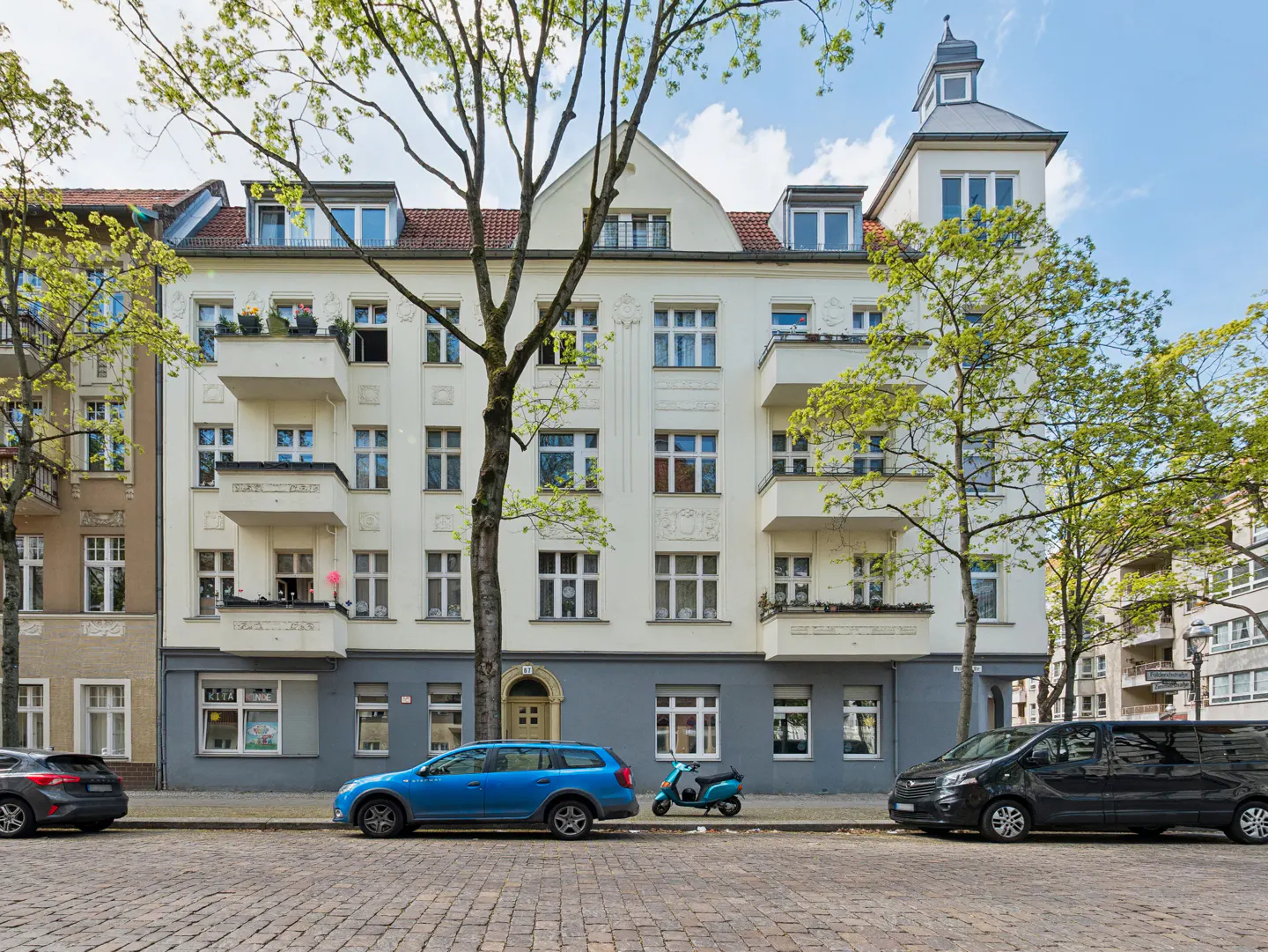 A cream-colored apartment building with balconies, cars parked on a cobblestone street.