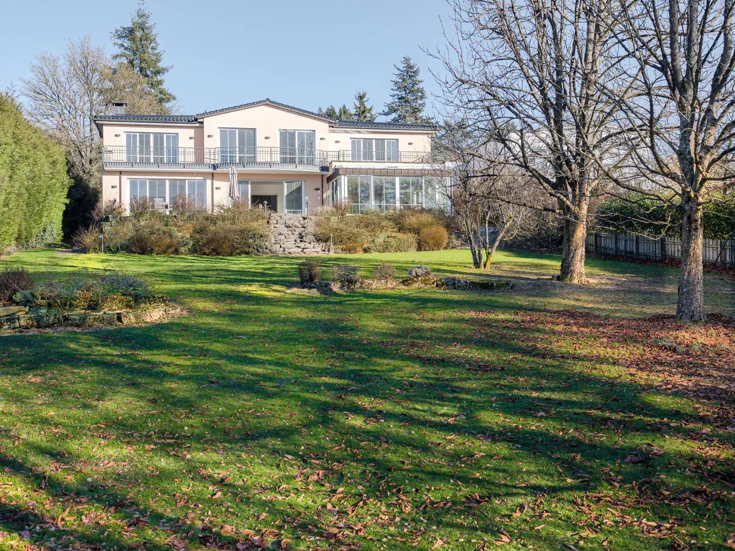Two-story peach house with balconies overlooks a large green lawn with trees and fallen leaves.