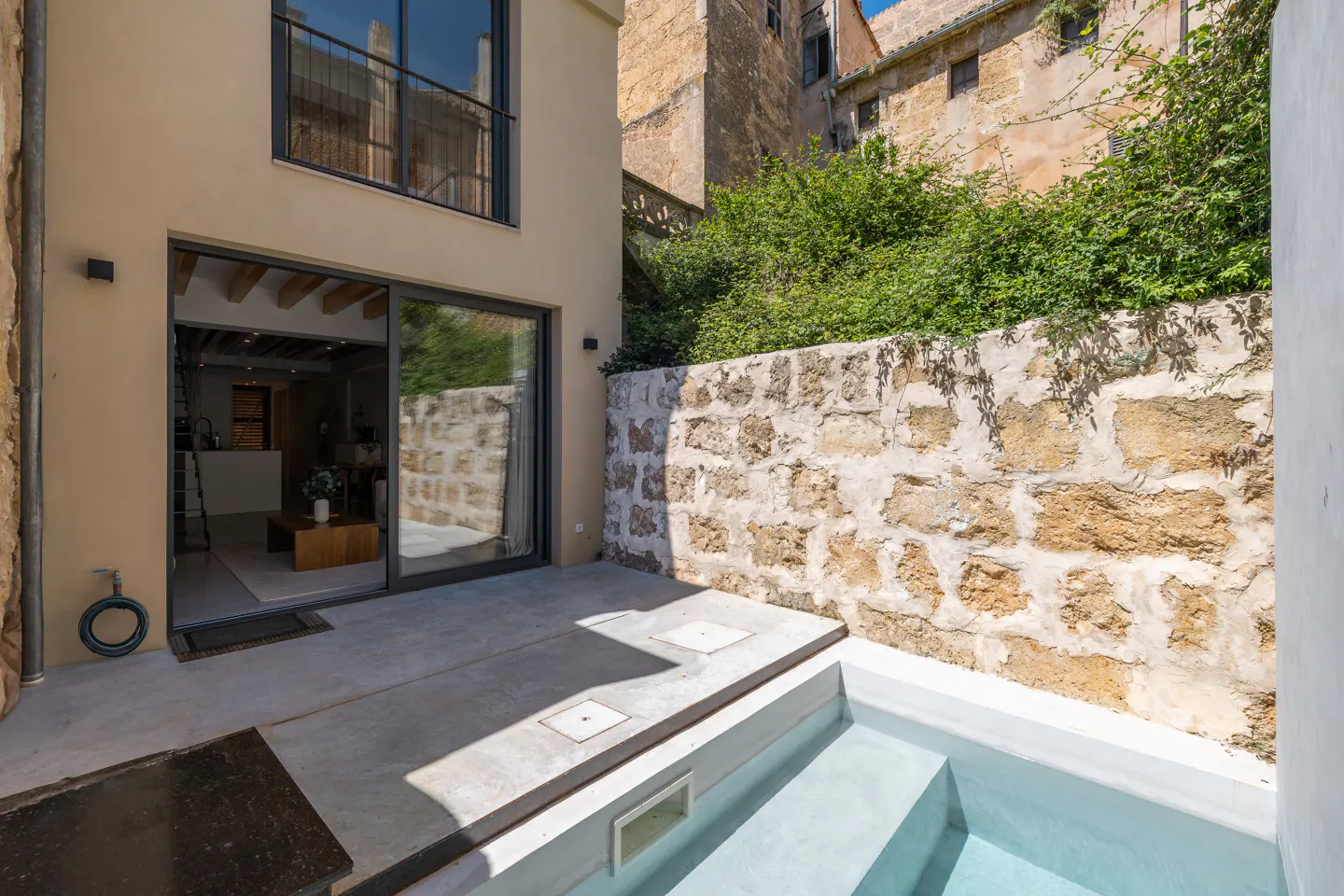 Exterior view of a modern home with a small pool. Stone wall with greenery, sliding glass doors, and a balcony are visible.