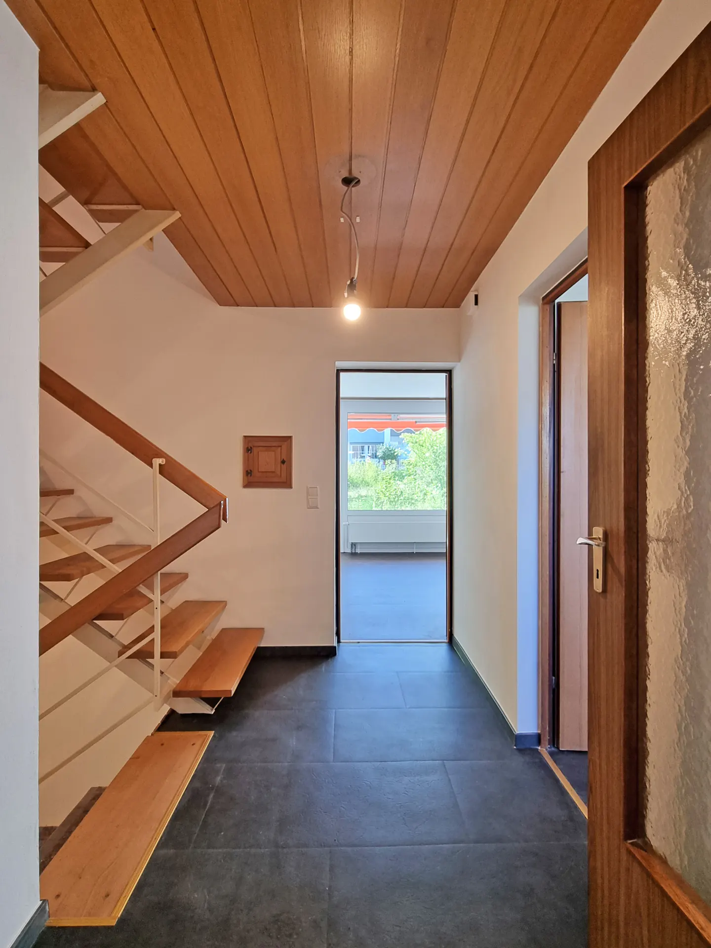 Interior view of a home's entryway with wood ceiling, stairs, and an open doorway to a bright room with a view.