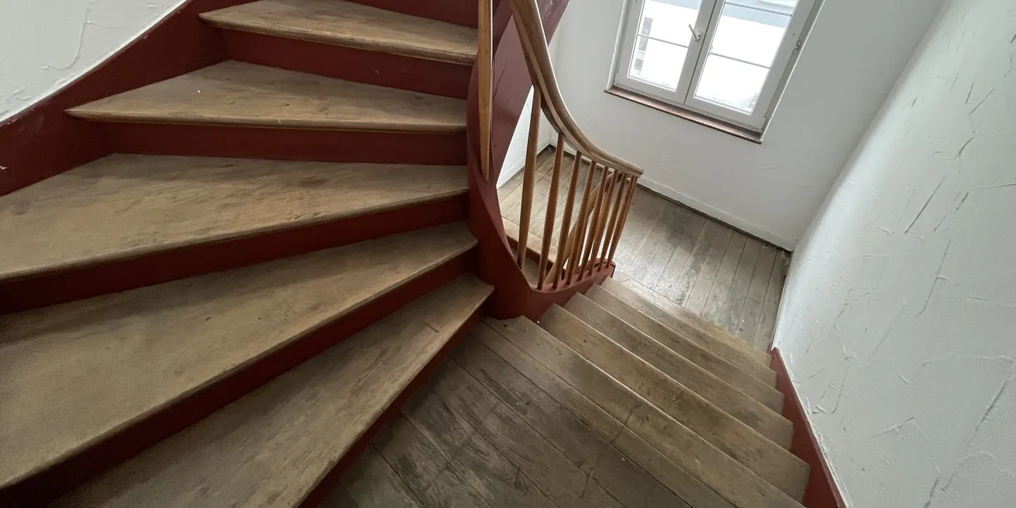 A high-angle shot of a wooden staircase with red trim and a curved banister, leading down to a wooden floor near a window.