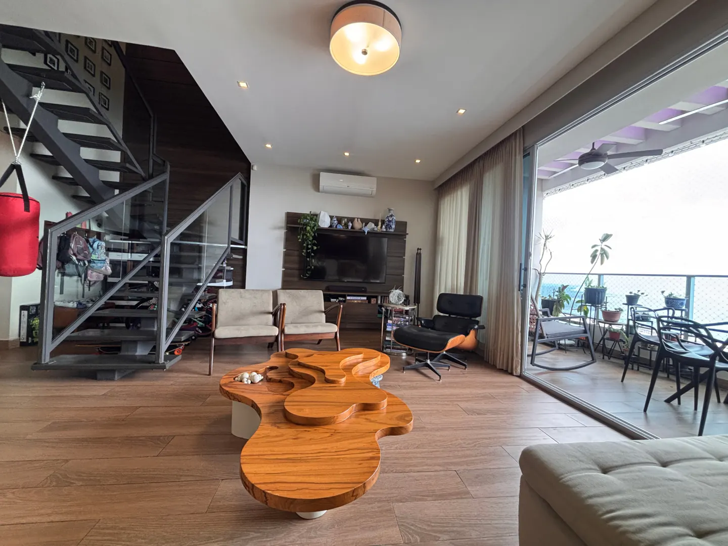 Bright living room with wood floors, a unique wood coffee table, and a balcony with ocean views. A metal staircase is on the left.