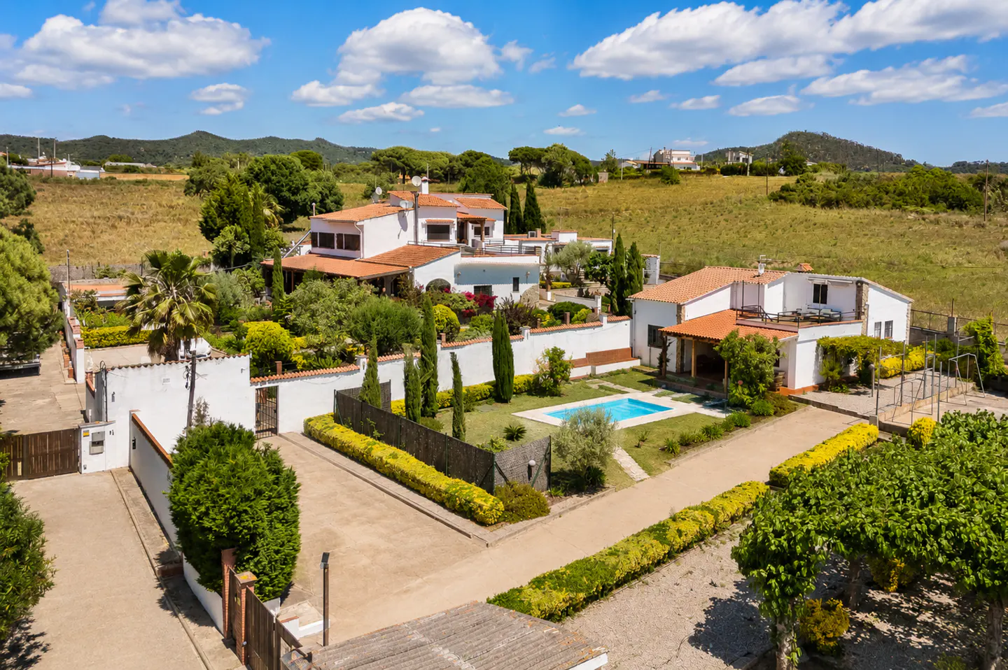 Aerial view of a white villa with a red tile roof, a pool, and lush green landscaping on a sunny day.