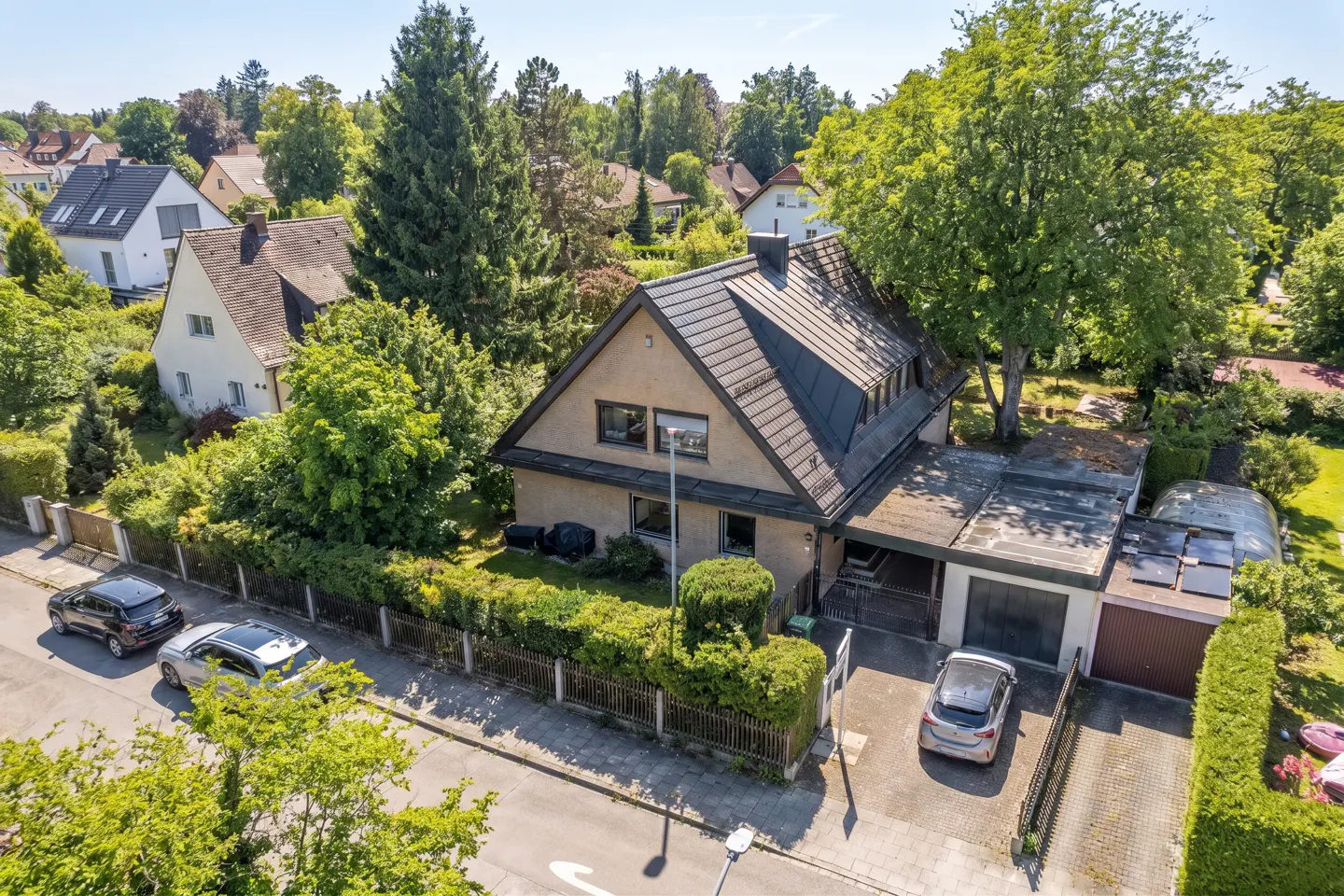 Aerial view of a tan house with a dark roof, surrounded by green trees and bushes, with cars parked nearby.