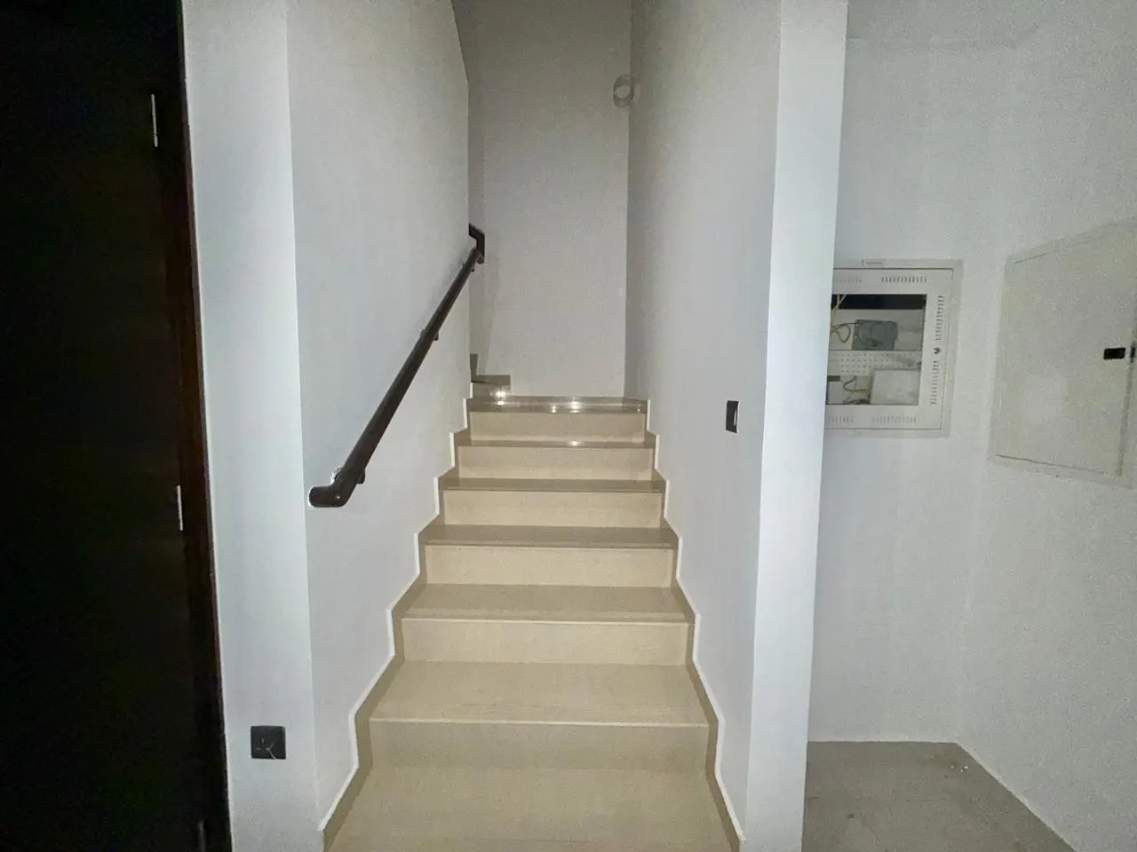 Interior view of a staircase with beige steps and a dark brown handrail against white walls. A dark door is visible on the left.