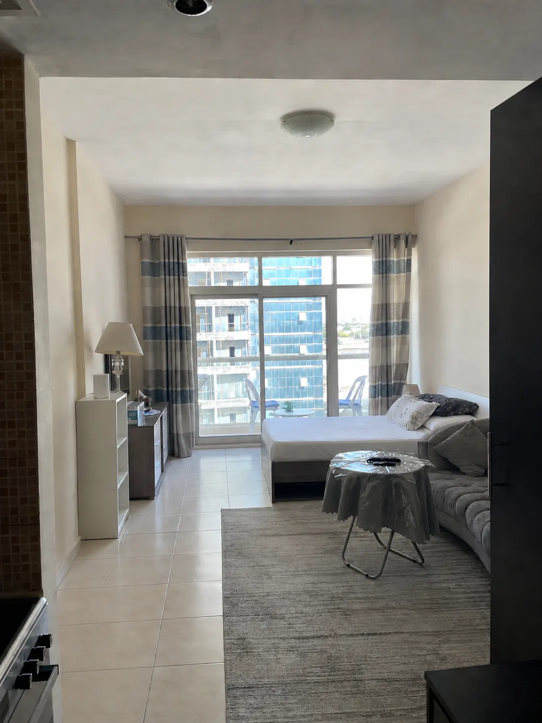Bright apartment interior with a bed, sofa, and table. Large windows offer a city view. Gray rug on tile floor. Blue and gray striped curtains.
