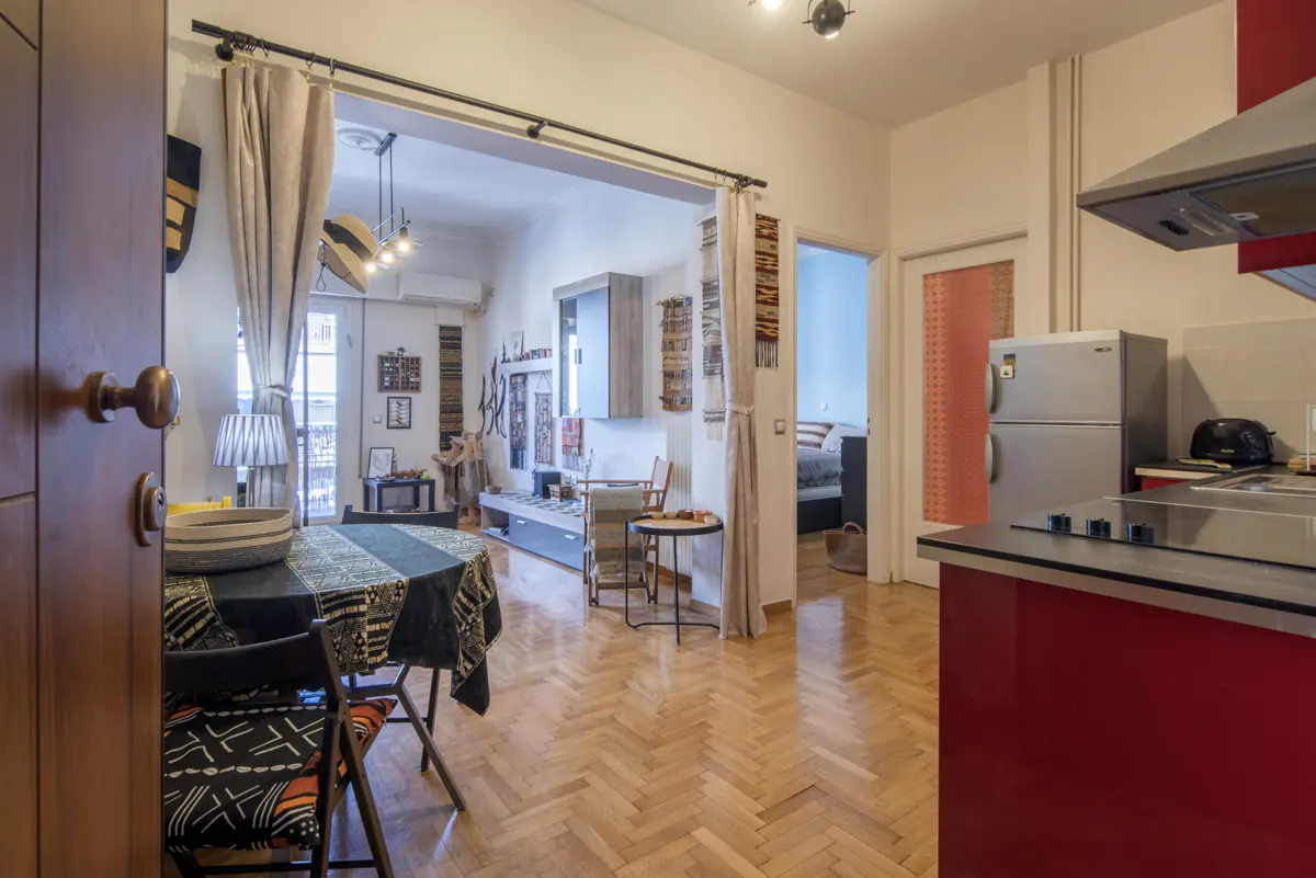 Open view of a modern apartment with a dining table, living room, and red kitchen cabinets. Hardwood floors and neutral walls create a bright, inviting space.