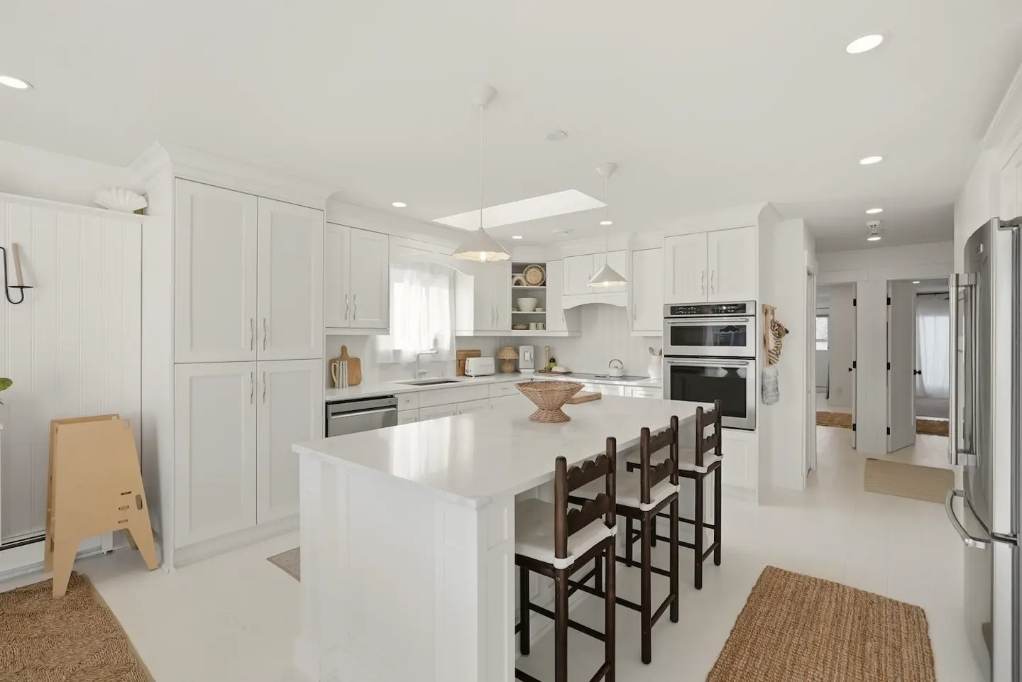 Bright, all-white kitchen with an island, dark wood stools, and stainless steel appliances.