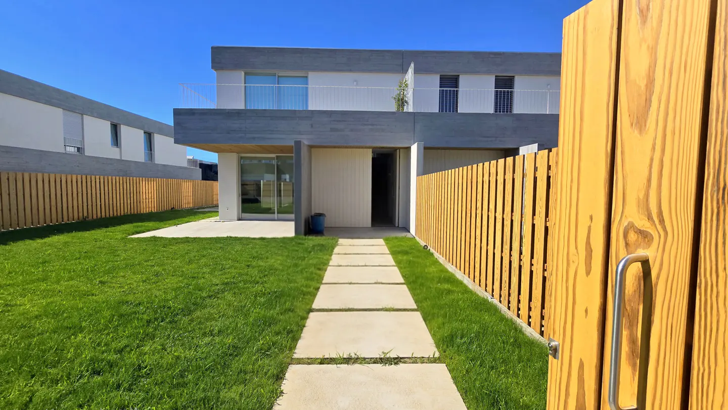 Modern two-story home with a green lawn, concrete walkway, and wooden privacy fence under a clear blue sky.