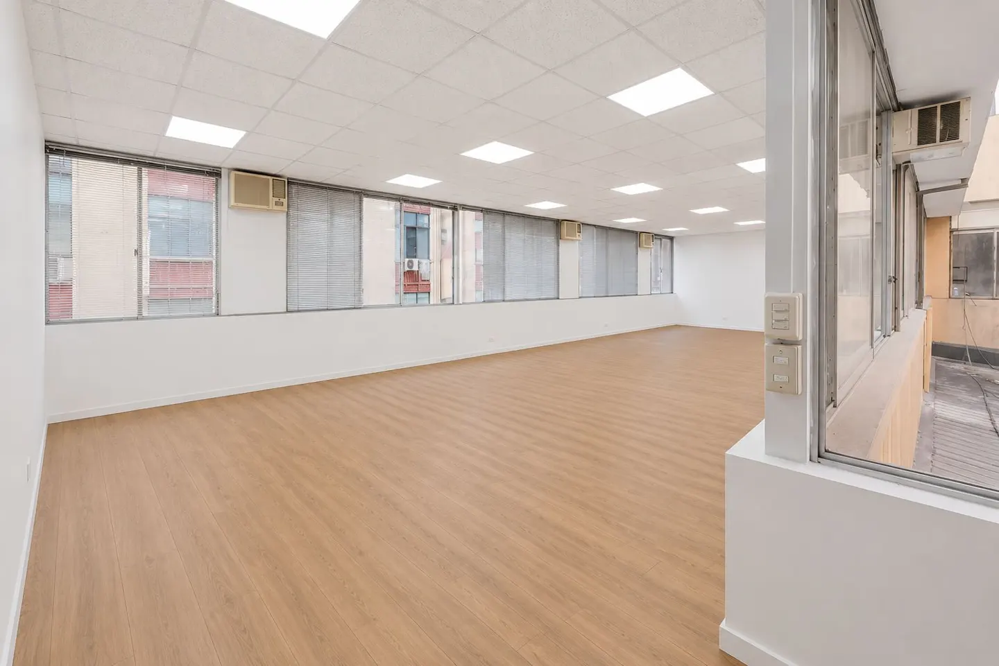 Bright, empty office space with wood floors, white walls, and large windows with gray blinds. Air conditioning units are visible above the windows.