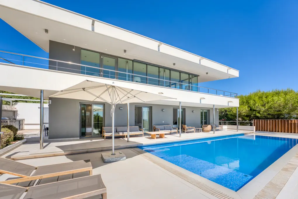 Two-story modern home with a pool. The house is gray and white with large glass windows. Lounge chairs and sofas surround the pool.