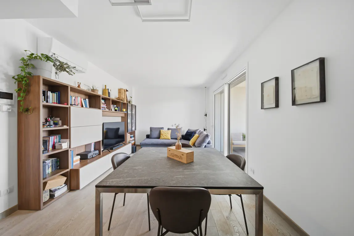Bright, modern living room with a gray dining table, brown chairs, and a large bookshelf with a TV. A gray sofa is visible in the background.