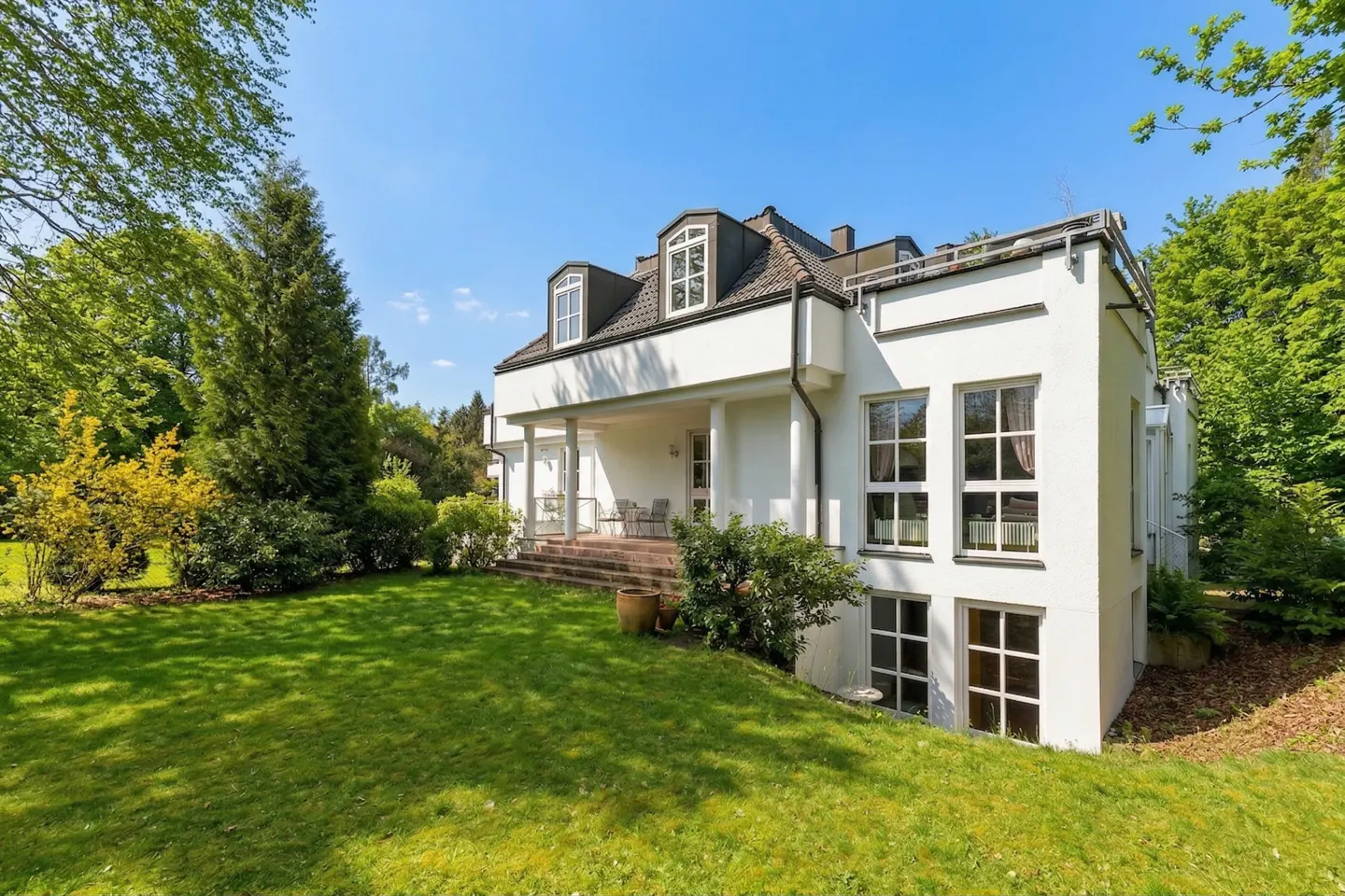 Exterior view of a white, two-story house with a green lawn and trees under a blue sky. The house has a covered porch with columns and large windows.
