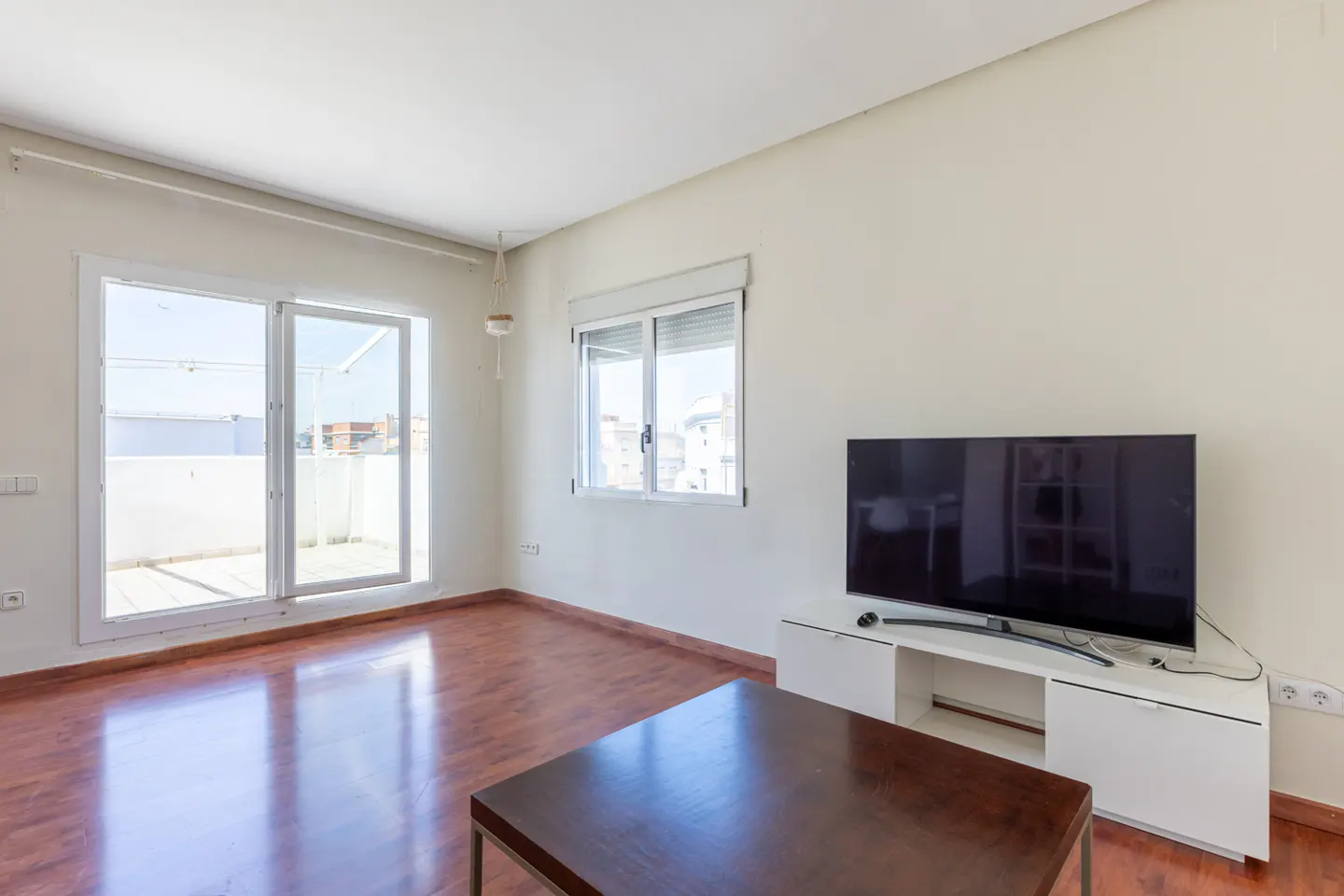 Bright living room with wood floors, white walls, and a large TV on a white stand. Sliding glass doors lead to a balcony.