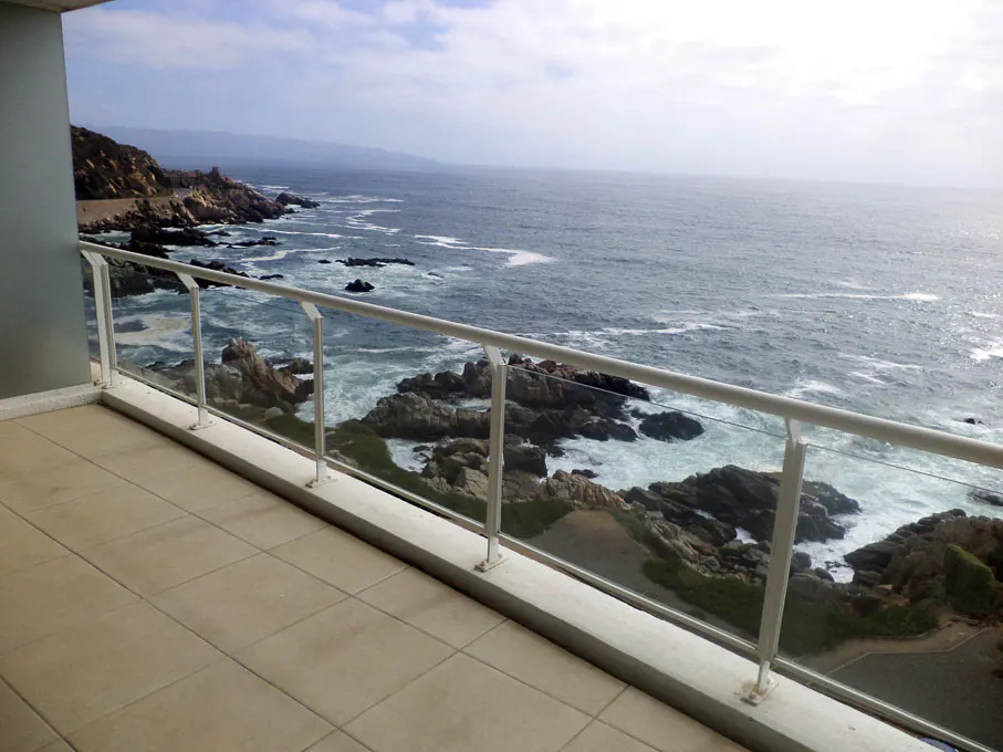 Balcony view of the ocean with rocks and waves. White railing and tan tile floor. Cloudy sky.