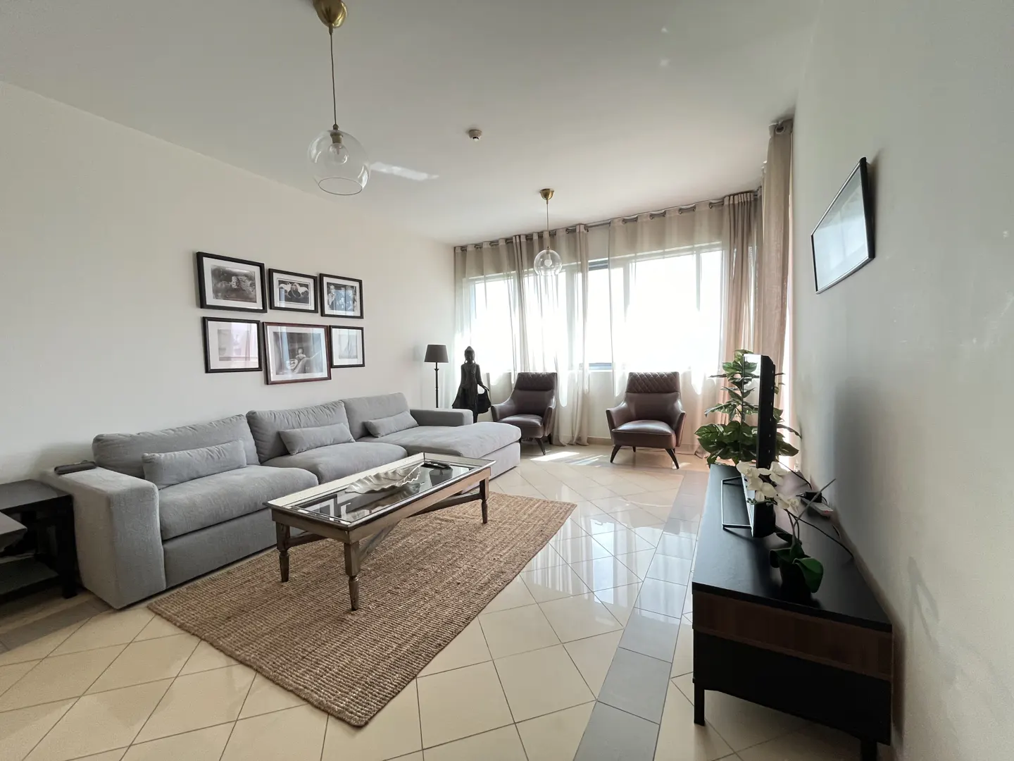 A bright living room with a gray sectional sofa, a glass coffee table on a jute rug, and two brown armchairs facing a large window.