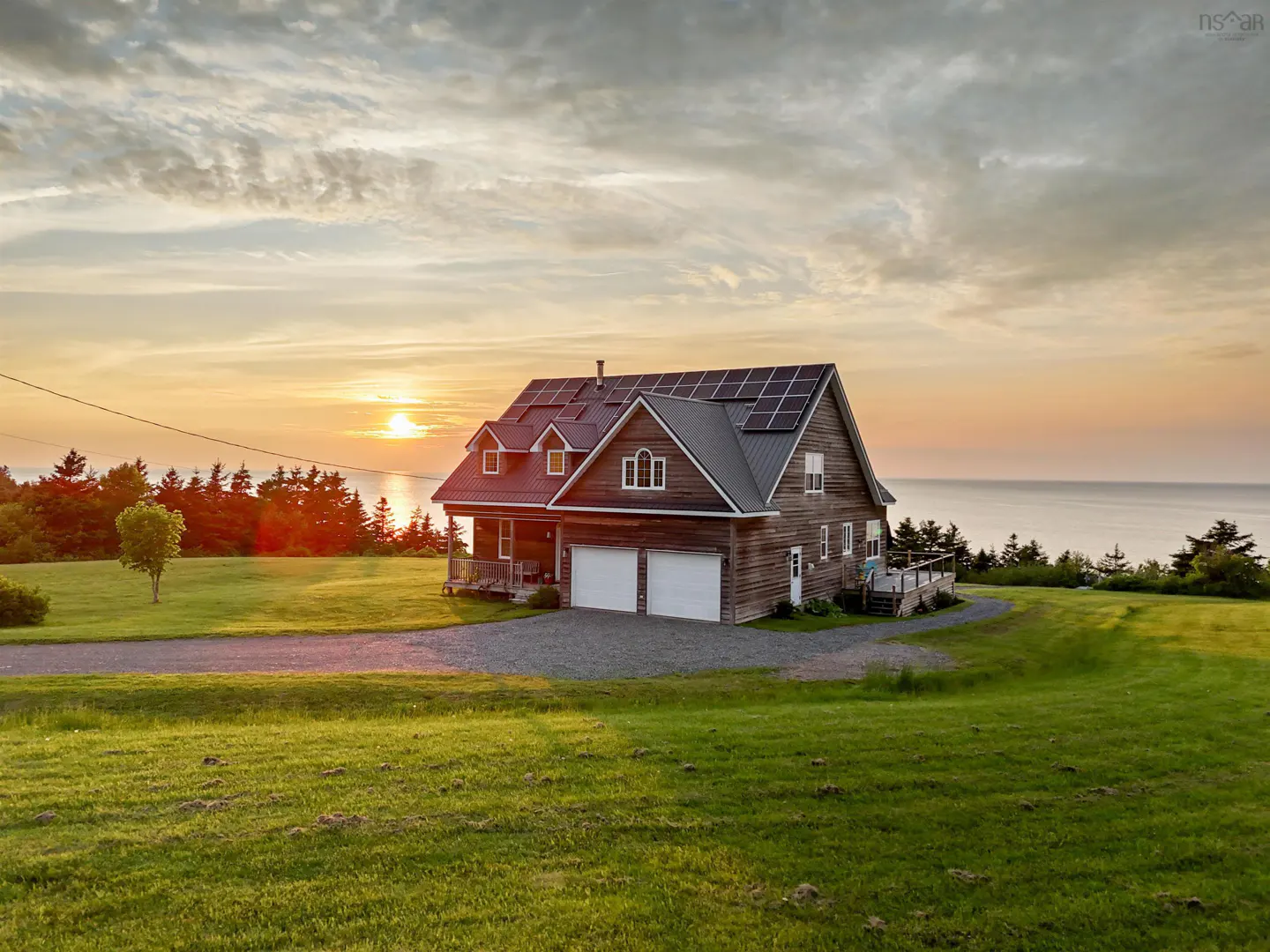 A two-story wood house with solar panels at sunset. The house has a two-car garage and is on a green lawn near the ocean.