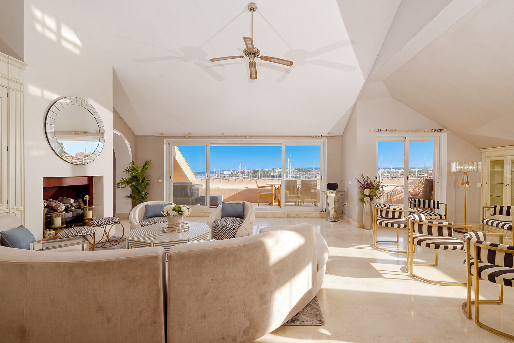 Bright, modern living room with a curved beige sofa, fireplace, and large glass doors opening to a sunny marina view. [AI-generated text]