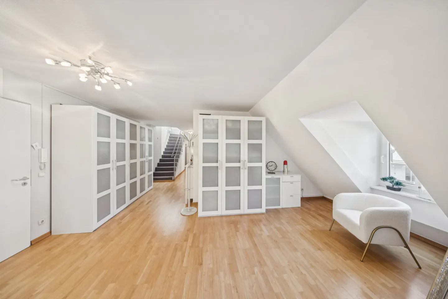 Bright attic room with wood floors, white walls, and built-in white cabinets. A white chair sits near a window with a plant. Stairs lead to another level.