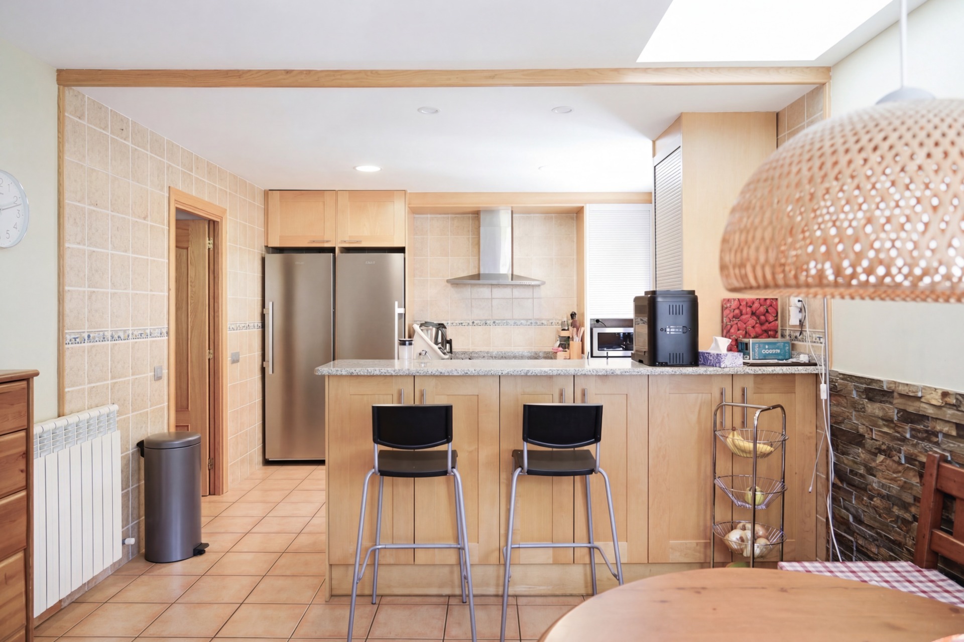 A bright kitchen with light wood cabinets, stainless steel appliances, and a granite countertop island with two black bar stools.