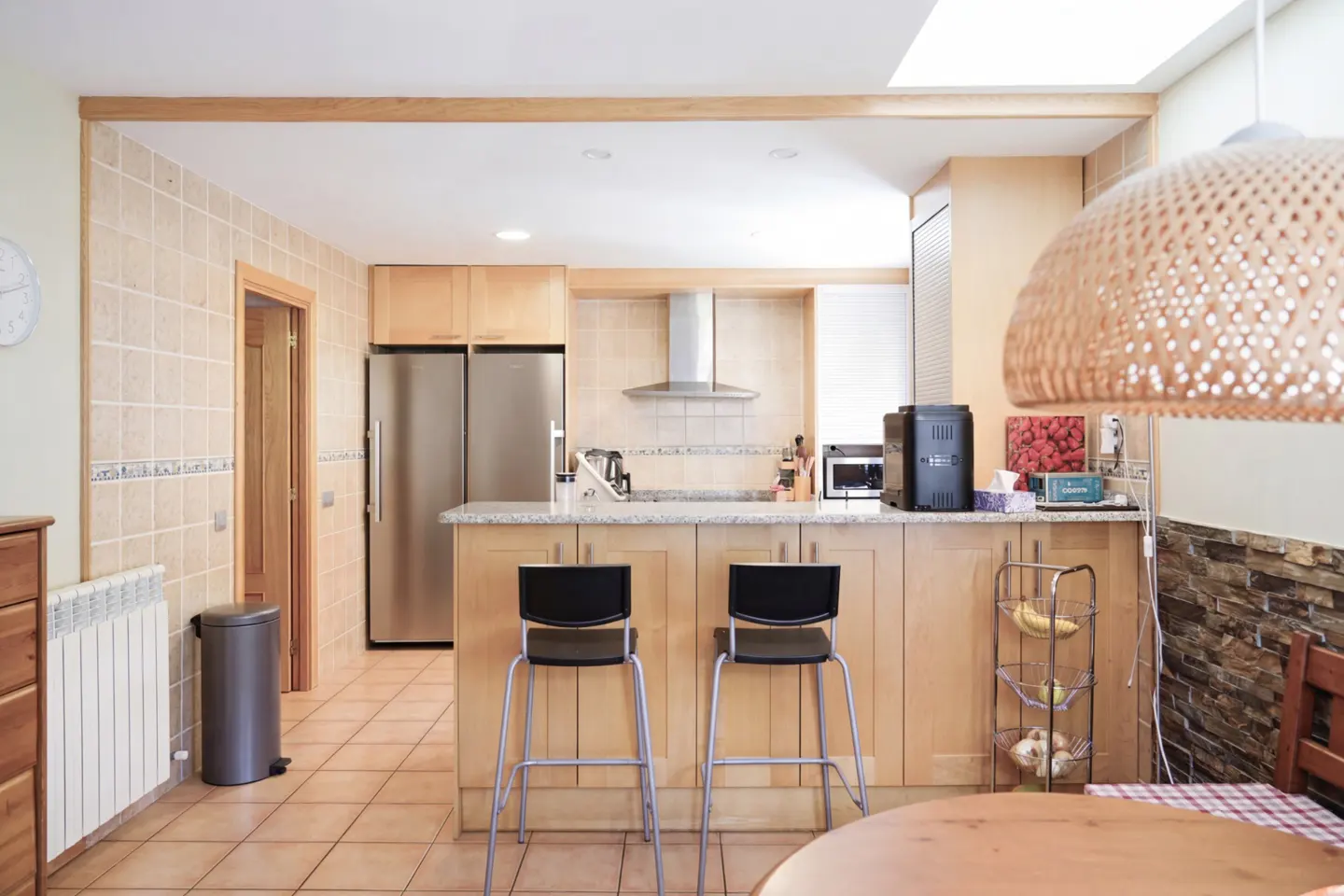 A bright kitchen with light wood cabinets, stainless steel appliances, and a granite countertop island with two black bar stools.