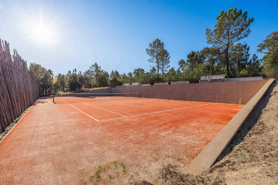 Outdoor tennis court with red clay surface, white lines, and net. Surrounded by trees and a wall under a clear blue sky.