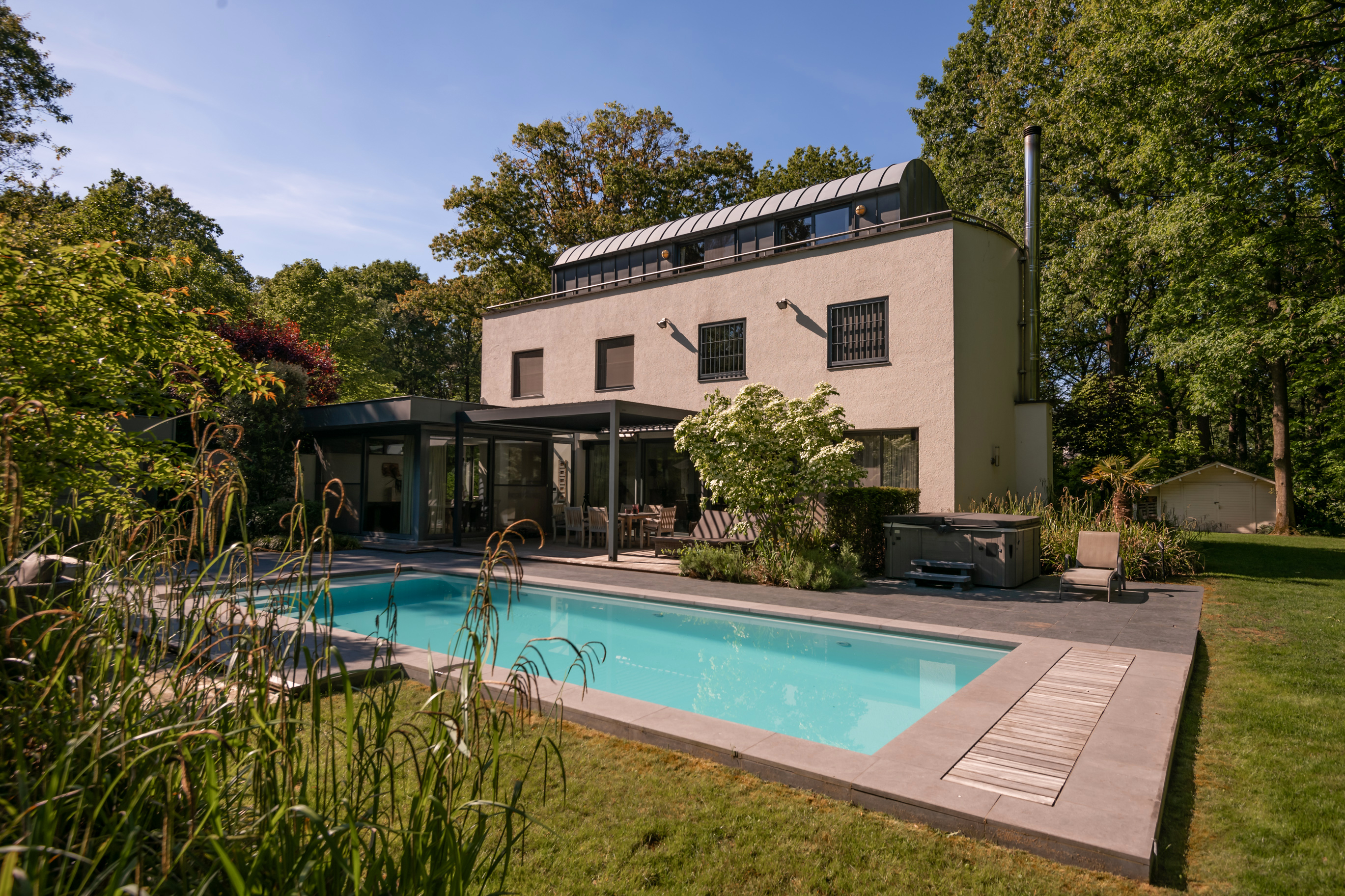 Modern beige house with a blue pool, patio, and green trees under a blue sky.