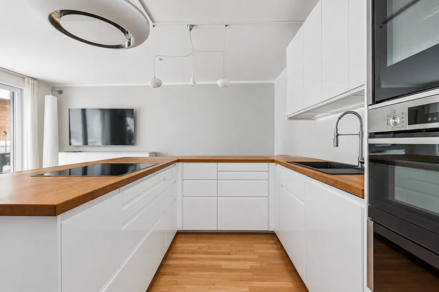 Modern kitchen with white cabinets, wooden countertops, built-in oven, and a view of a living area with a wall-mounted TV.