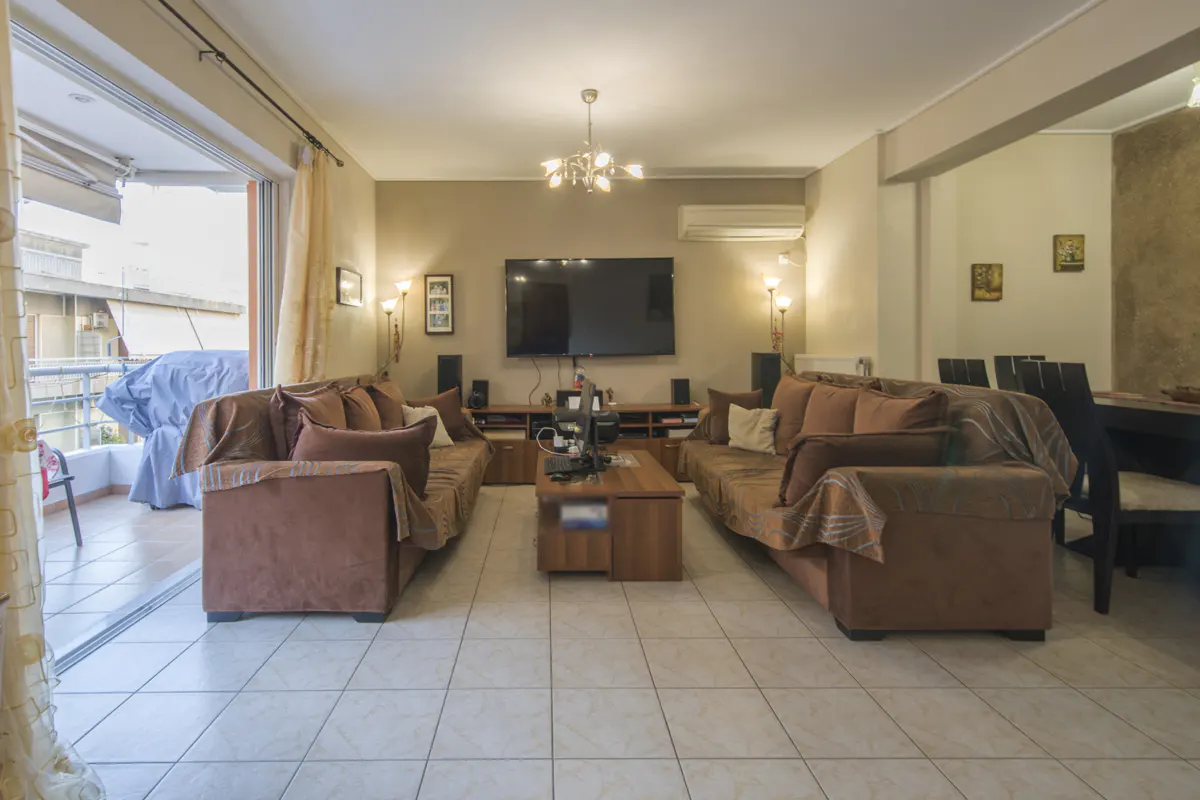 Living room with brown sofas, a TV, and a balcony. The room has tile floors and neutral-colored walls.