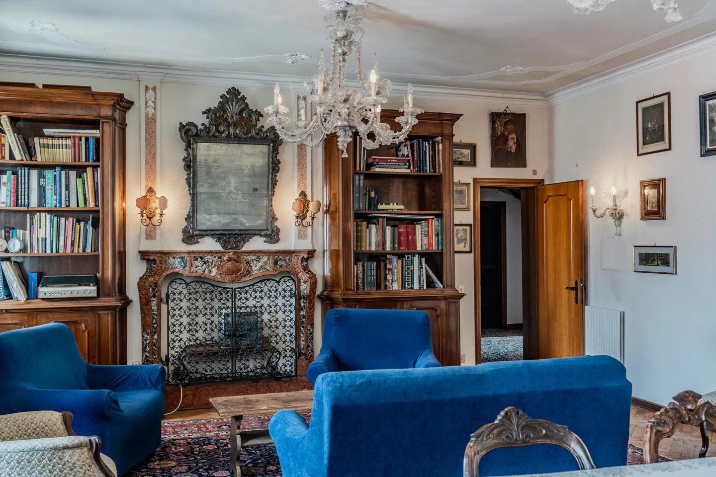 Elegant living room with blue sofas, a marble fireplace, crystal chandelier, and floor-to-ceiling wooden bookshelves.