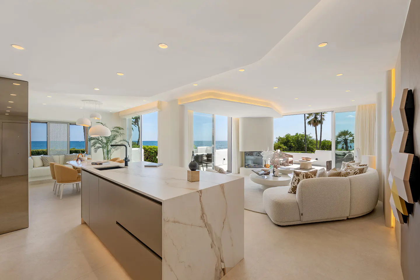 Modern open-plan living area with a marble kitchen island, curved white sofa, and floor-to-ceiling windows overlooking ocean.