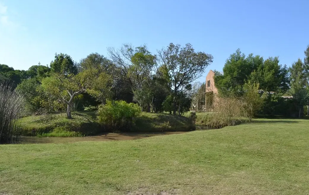 Lush green lawn with a stream, trees, and a brick house in the background under a clear blue sky.