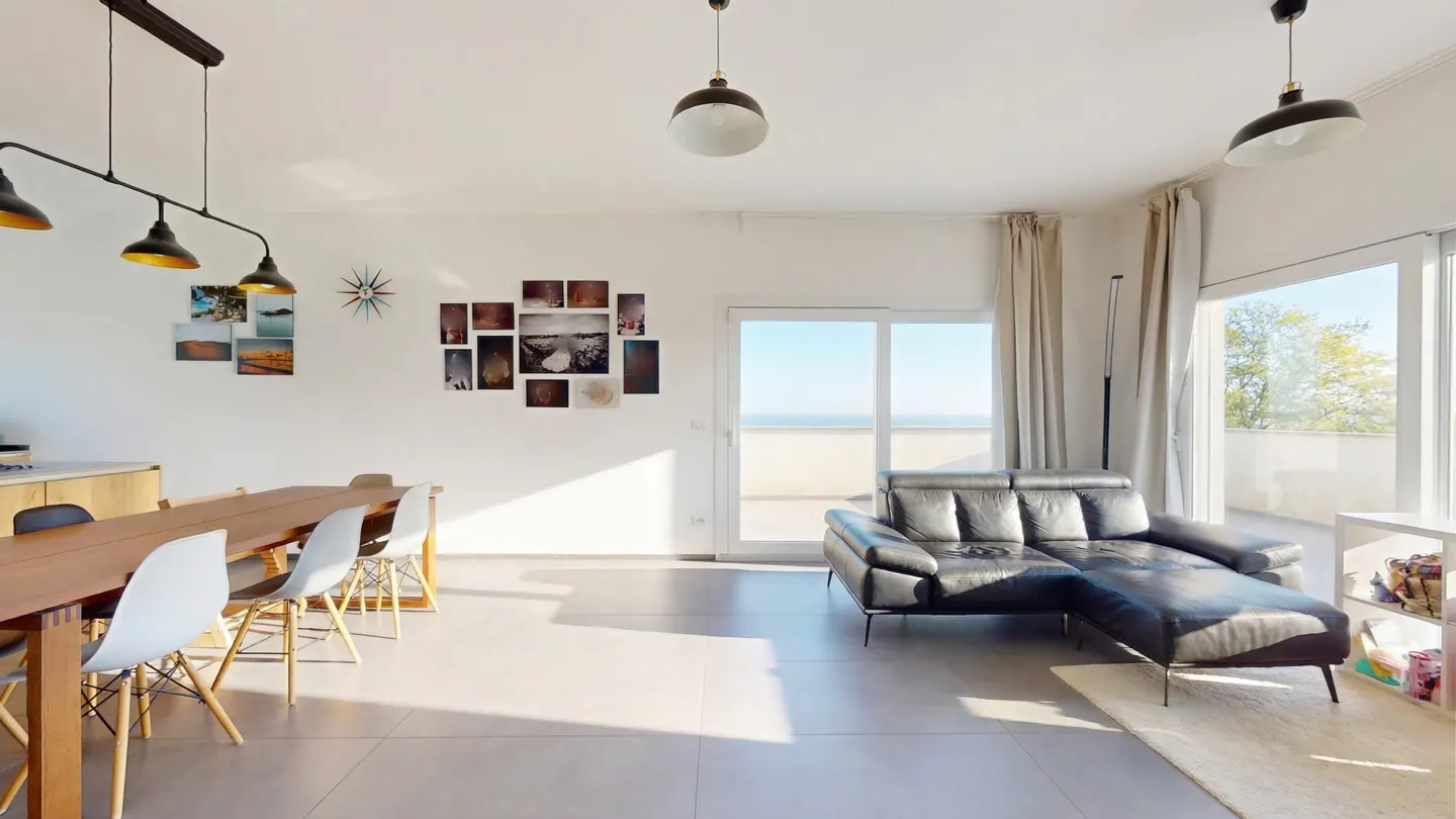 Bright, open-concept living space with a dining table, black leather sofa, and ocean view through sliding glass doors.
