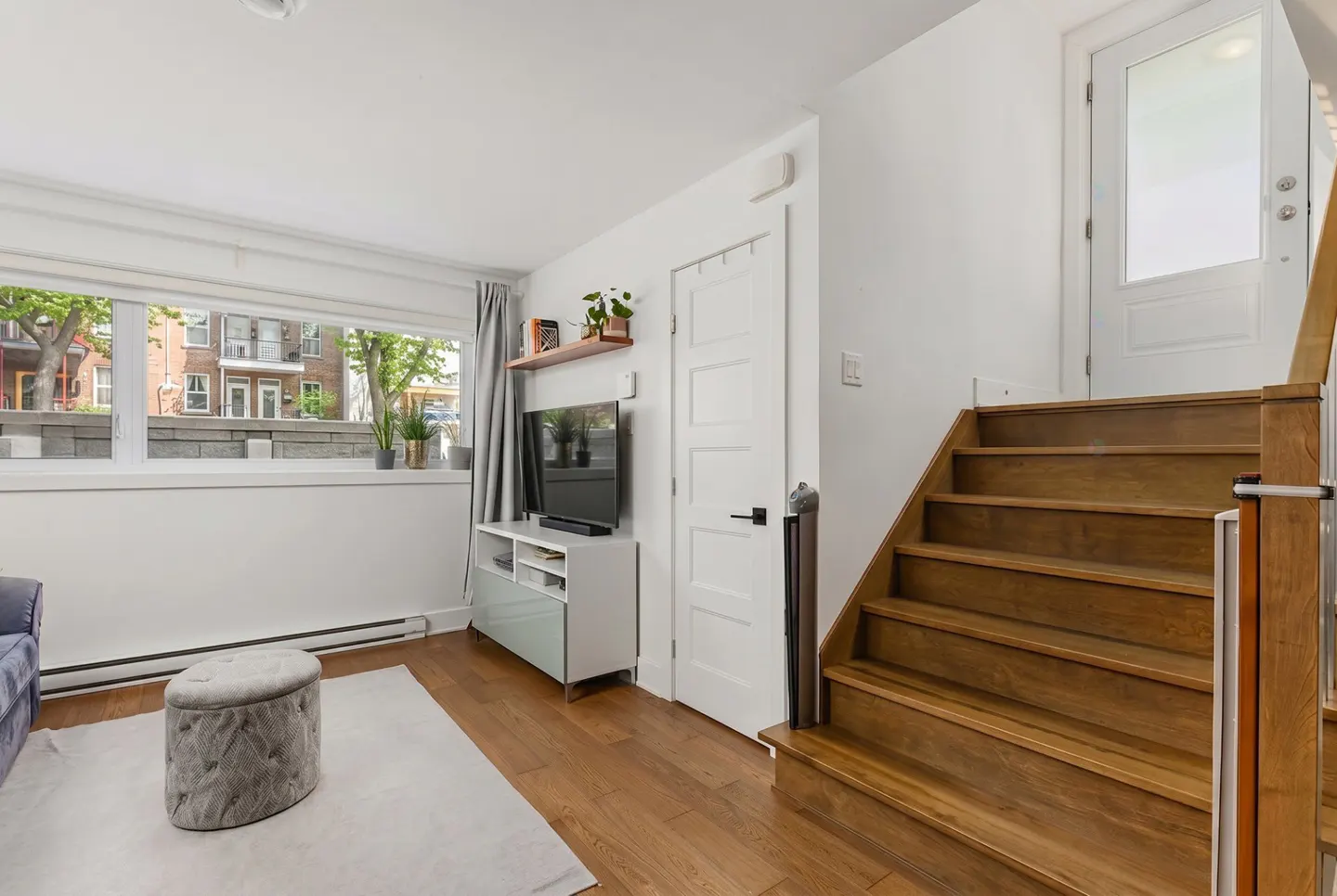 Bright living room with hardwood floors, white walls, and a large window. Stairs lead to a white door with a glass panel. A TV sits on a white cabinet.