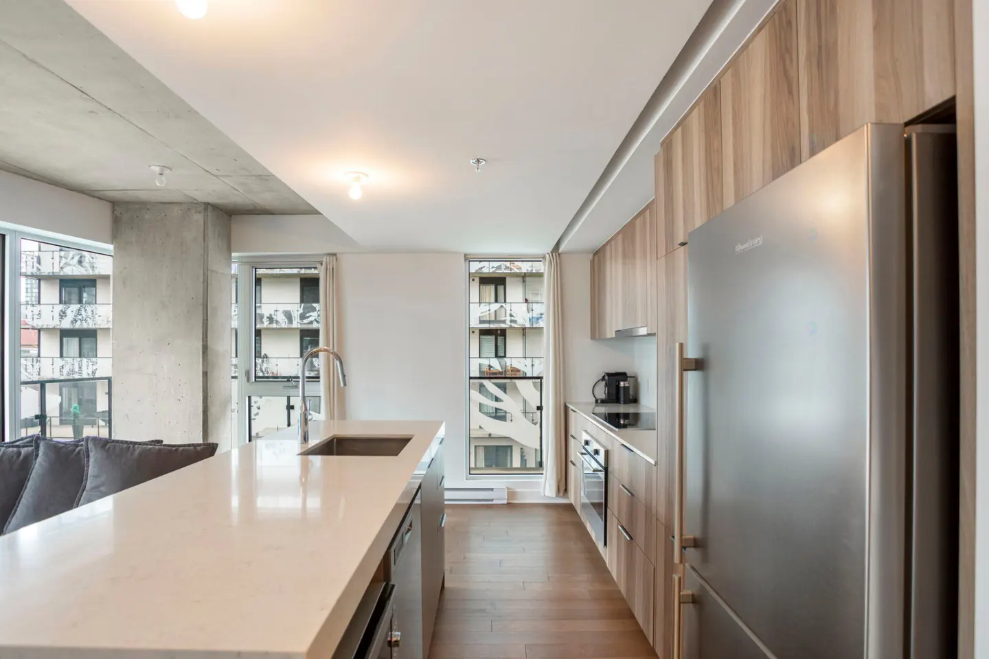 Modern kitchen with a white quartz island, stainless steel refrigerator, wood cabinets, and large windows with city views.