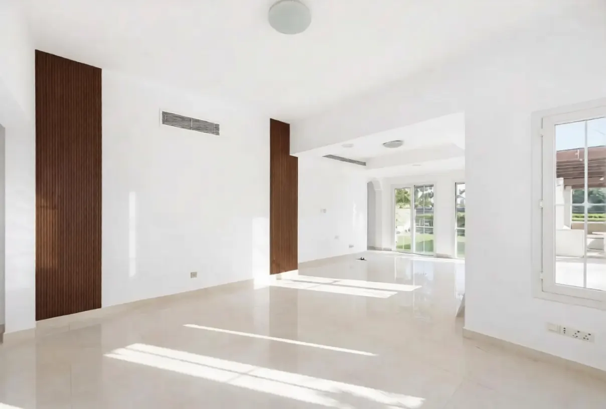 Bright, empty room with white walls, light tile floor, and brown wood accents. A window shows an outdoor patio.