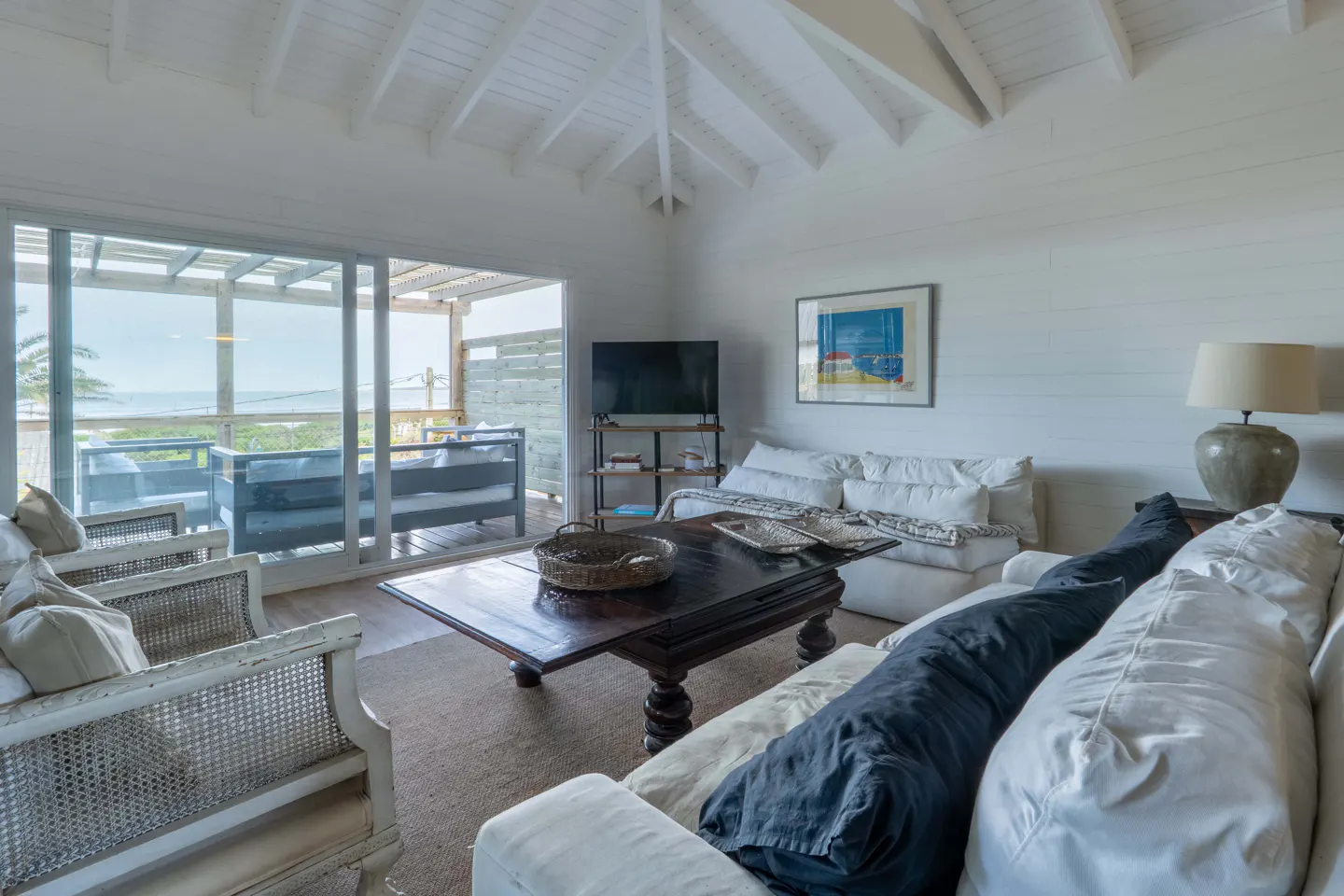Bright living room with white walls, ceiling, and furniture. A dark wood coffee table sits on a tan rug. Sliding glass doors lead to a deck with ocean views.