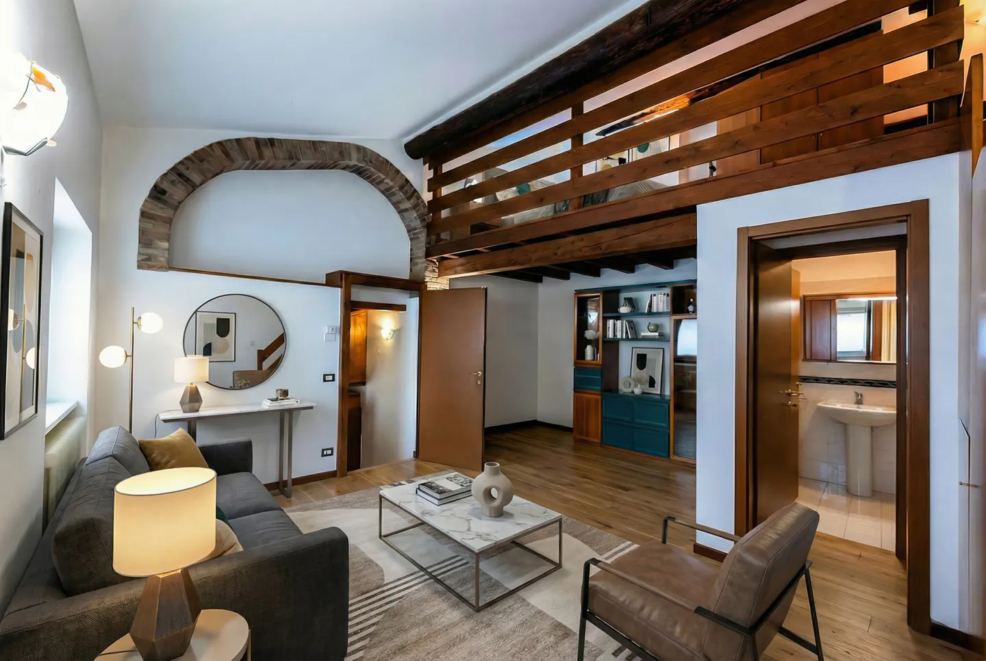 Living room with gray sofa, marble coffee table, and brown leather chair. Wooden beams on ceiling and a brick archway. Doorway to bathroom.
