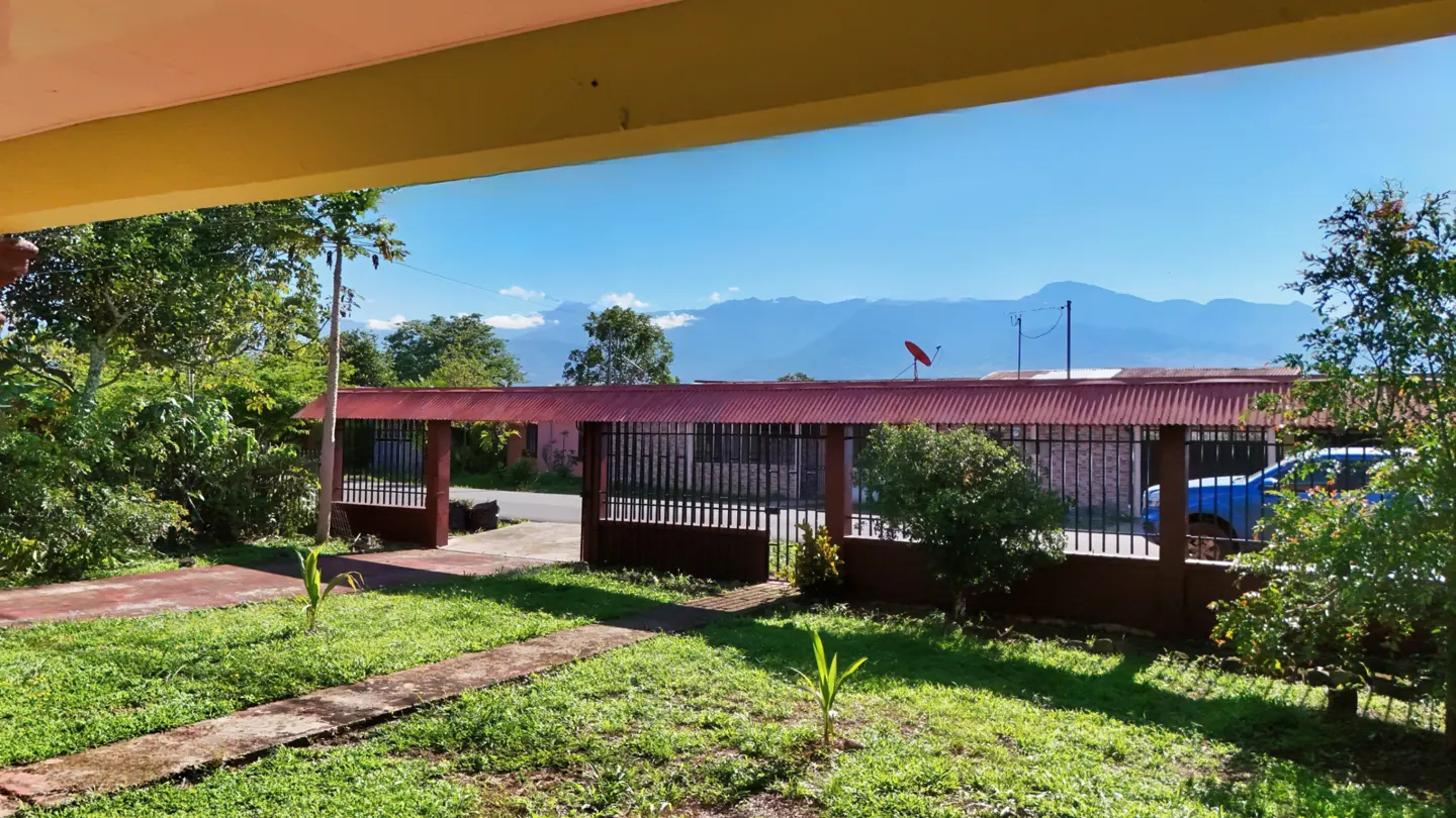View from a porch of a home's front yard with a red metal gate, green lawn, and mountains in the distance under a blue sky.