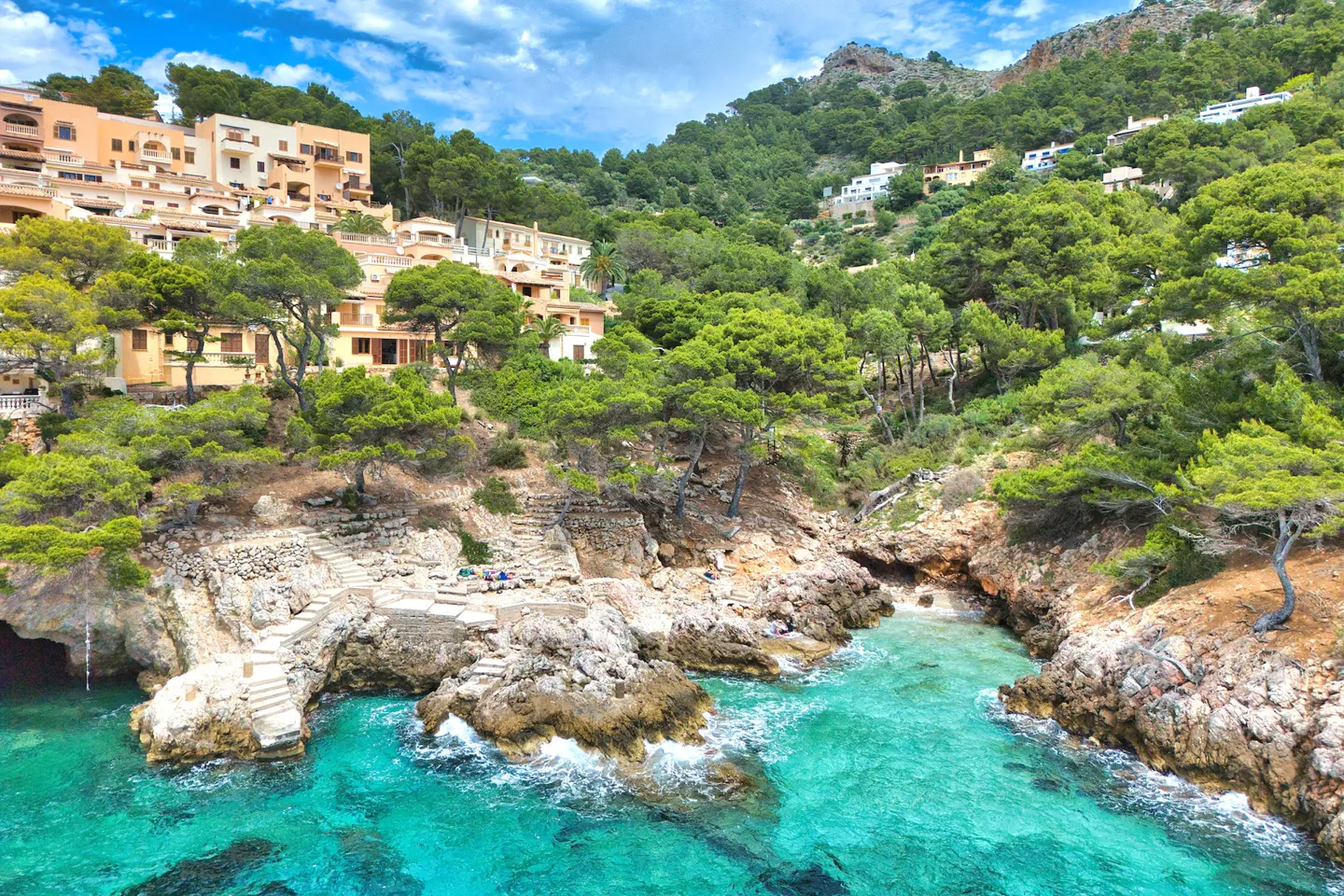 Coastal view of tan buildings on a hill, green trees, and turquoise water with rocky shoreline.