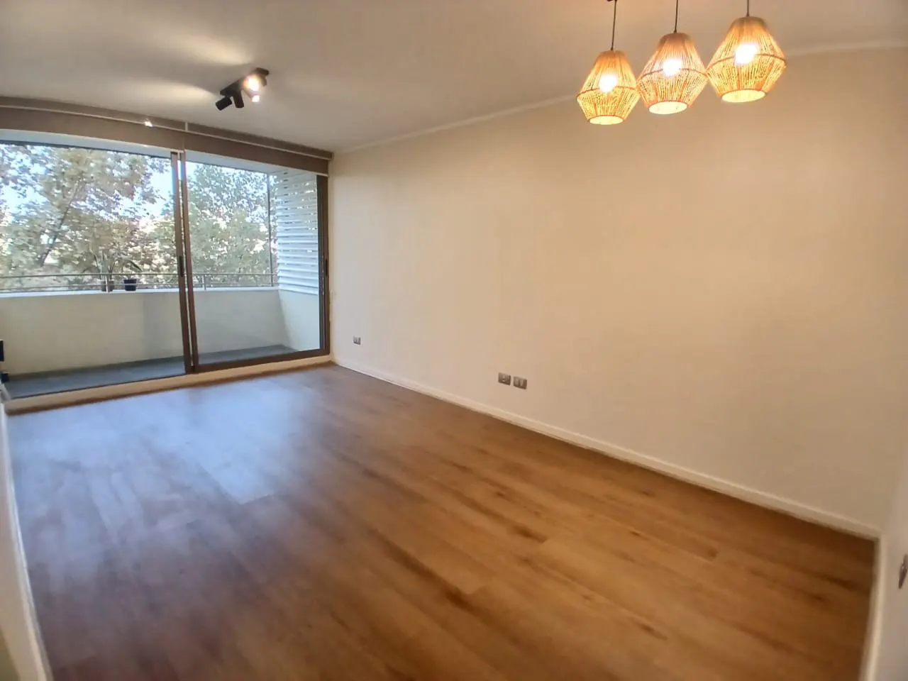 Empty living room with wood floors, white walls, and a balcony with trees visible through the glass doors. Three pendant lights hang from the ceiling.