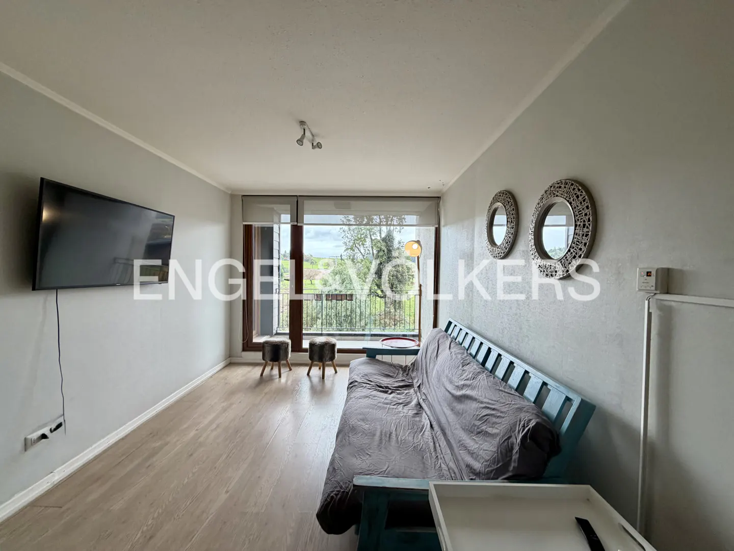 Living room with a blue futon, TV, and two round mirrors. A large window shows a green landscape. Engel & Völkers logo is overlaid.