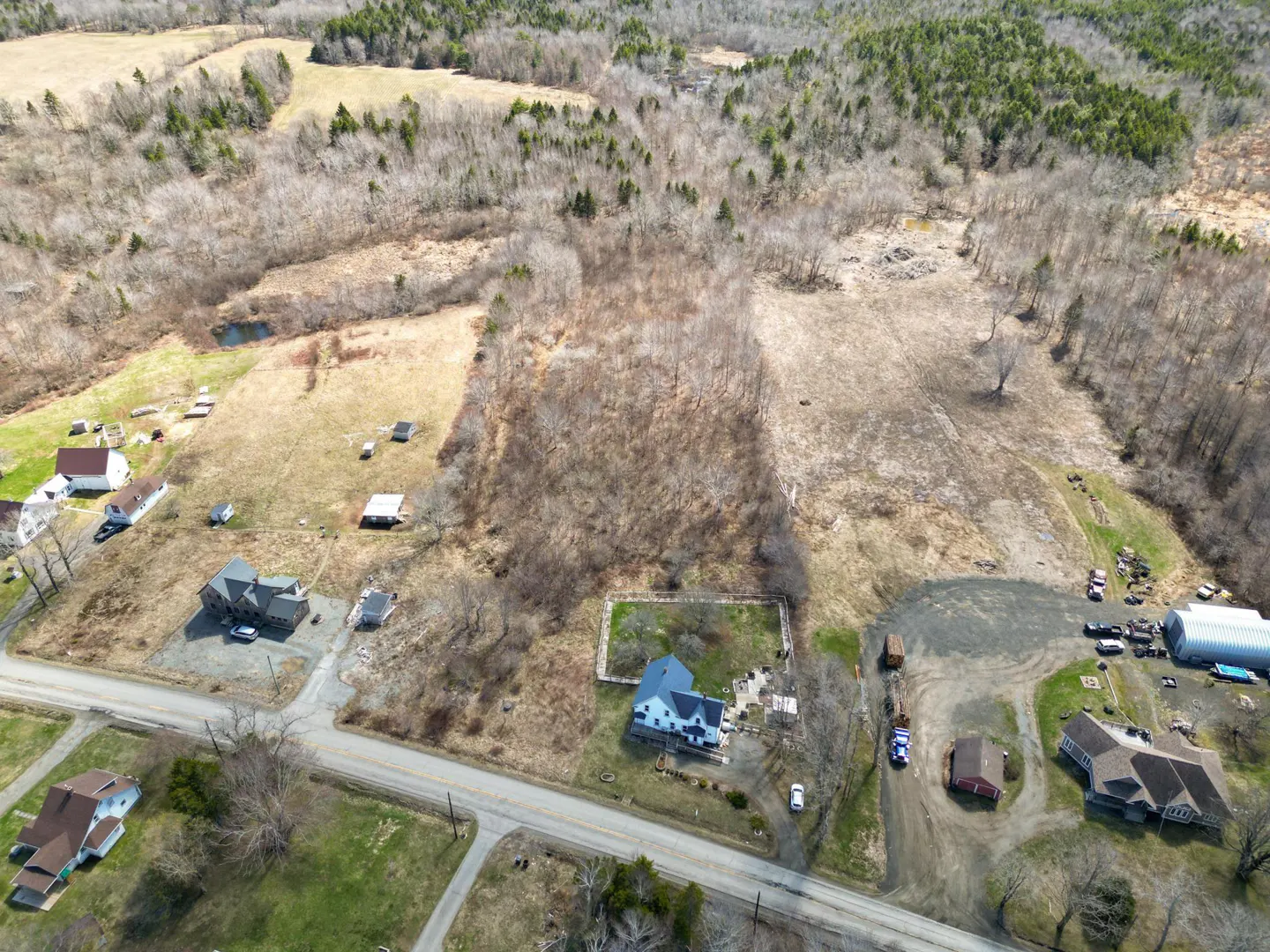 Aerial view of a rural landscape with houses, fields, and a forest in the background. A road runs through the scene.