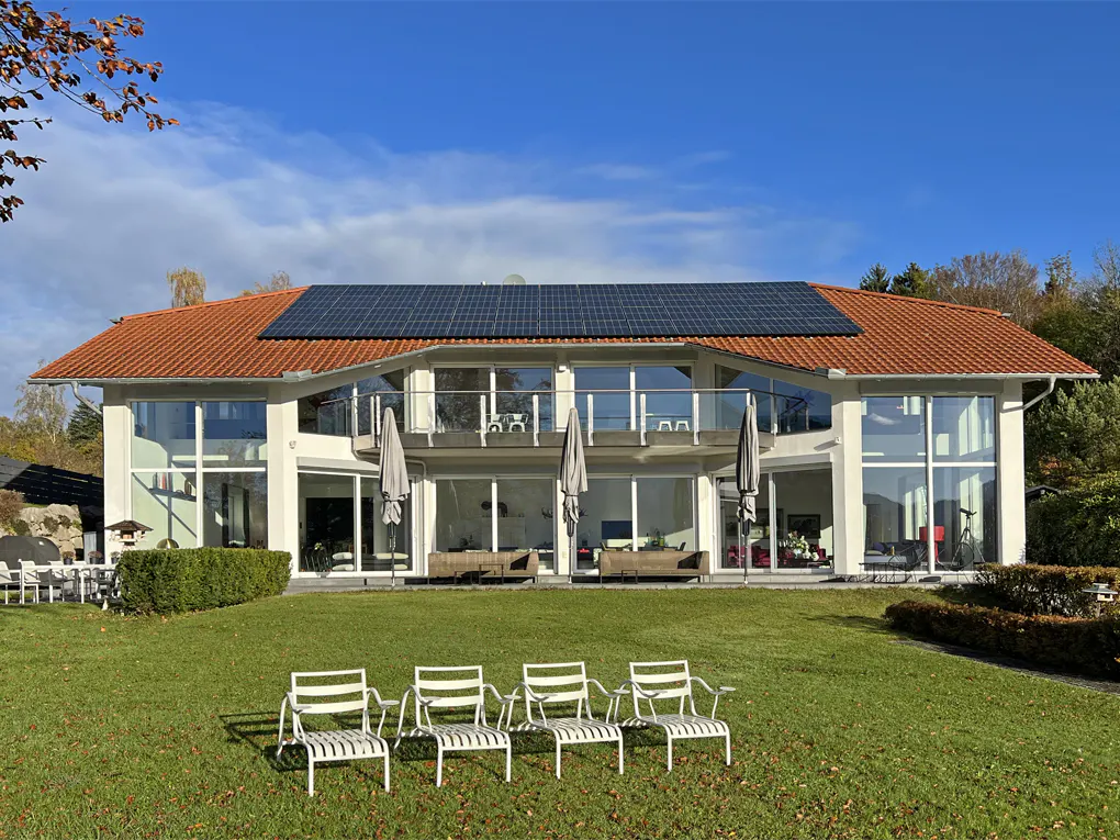 Modern white house with a red tile roof and solar panels, overlooking a green lawn with white chairs.