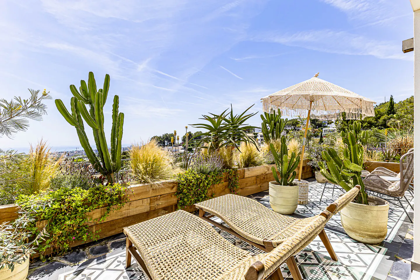 Outdoor patio with two woven lounge chairs, a fringed umbrella, and potted cacti against a blue sky.