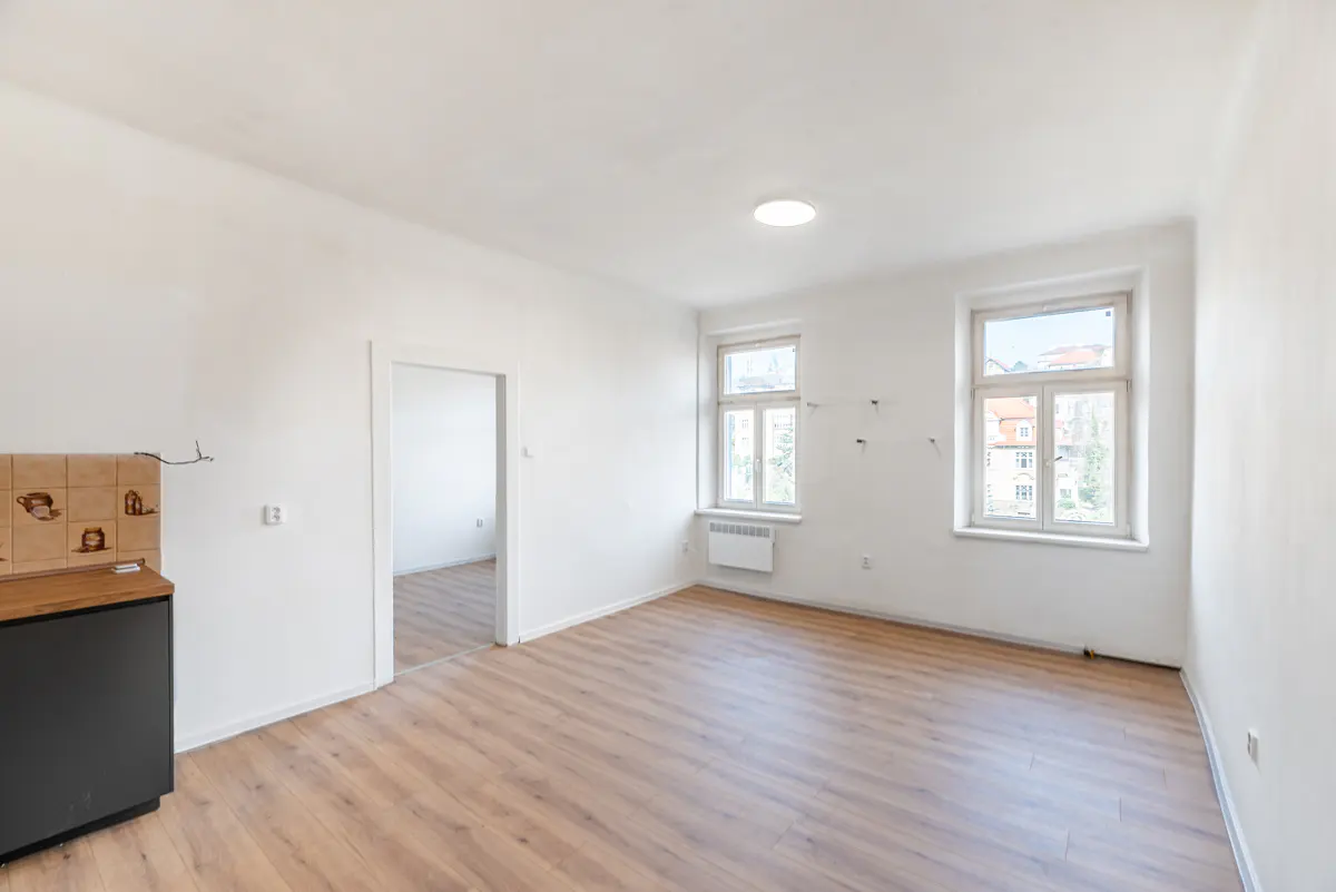 Bright, empty room with light wood floors, white walls, and two windows. A doorway leads to another room. A black cabinet is on the left.